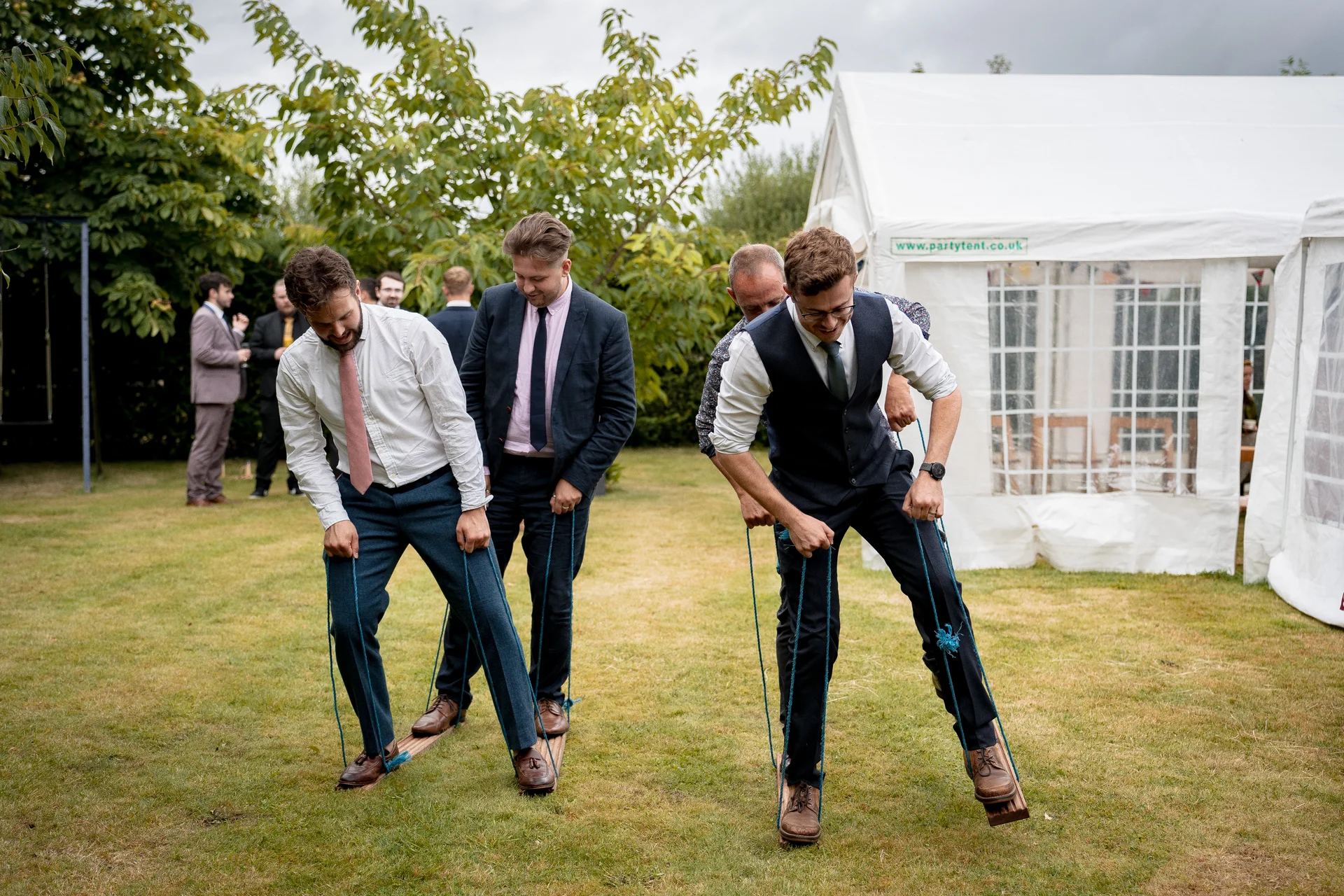 Groomsmen participate in a fun lawn game outside a white marquee tent during a wedding reception.