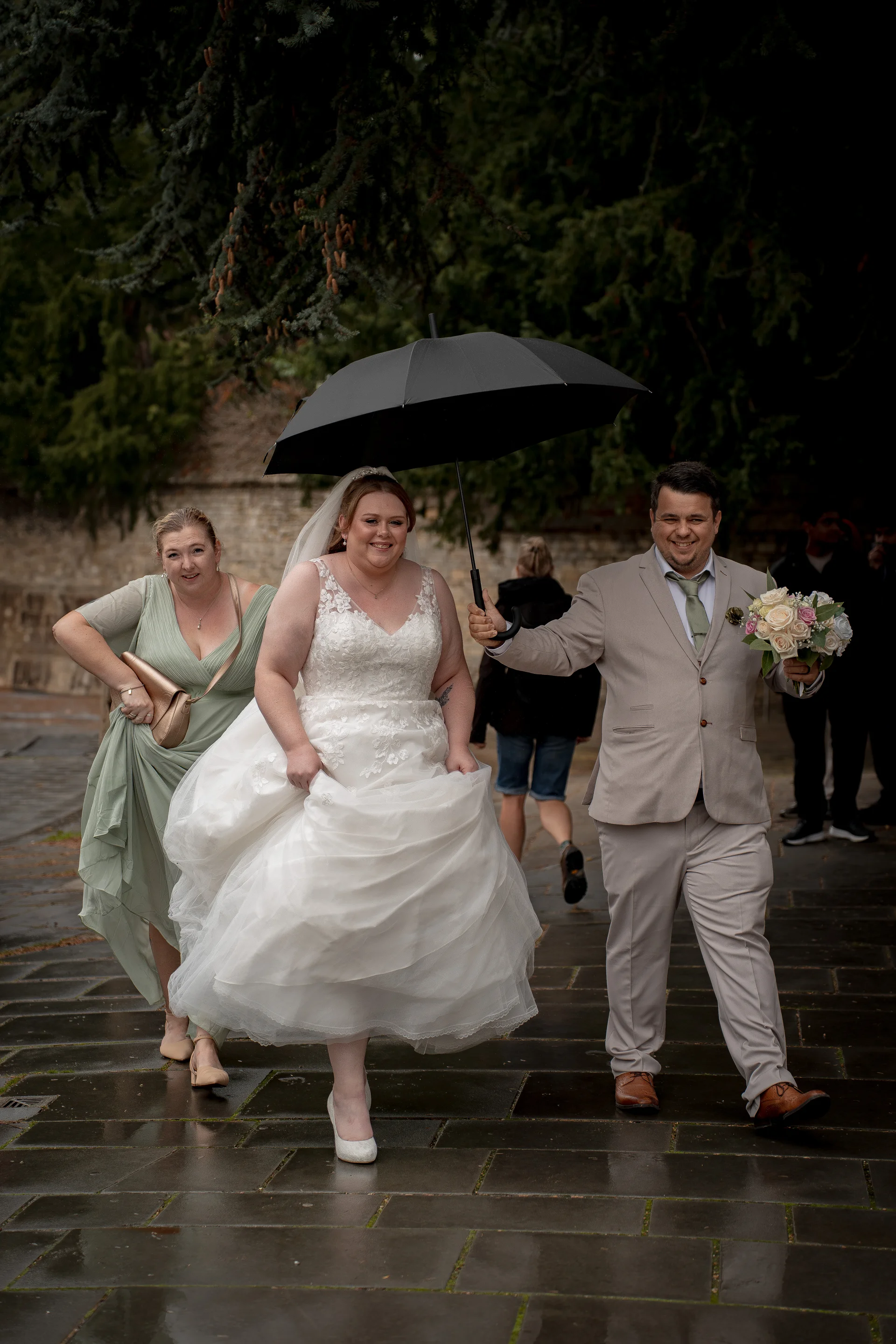 A smiling bride holding her dress and a groom holding an umbrella walk alongside a bridesmaid on a wet paved path outdoors.