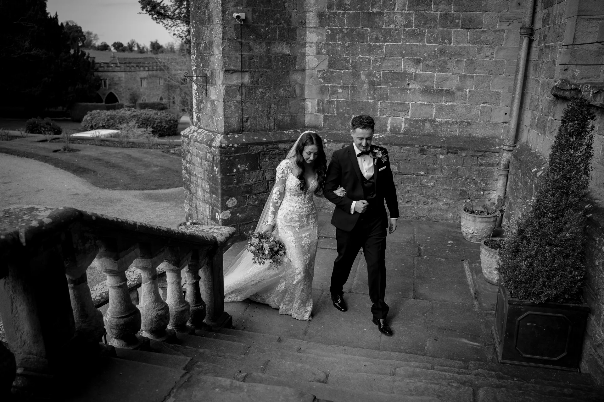 A newlywed couple walks down grand stone steps outside a historic stone venue, with the bride holding a bouquet and wearing a lace gown and veil.