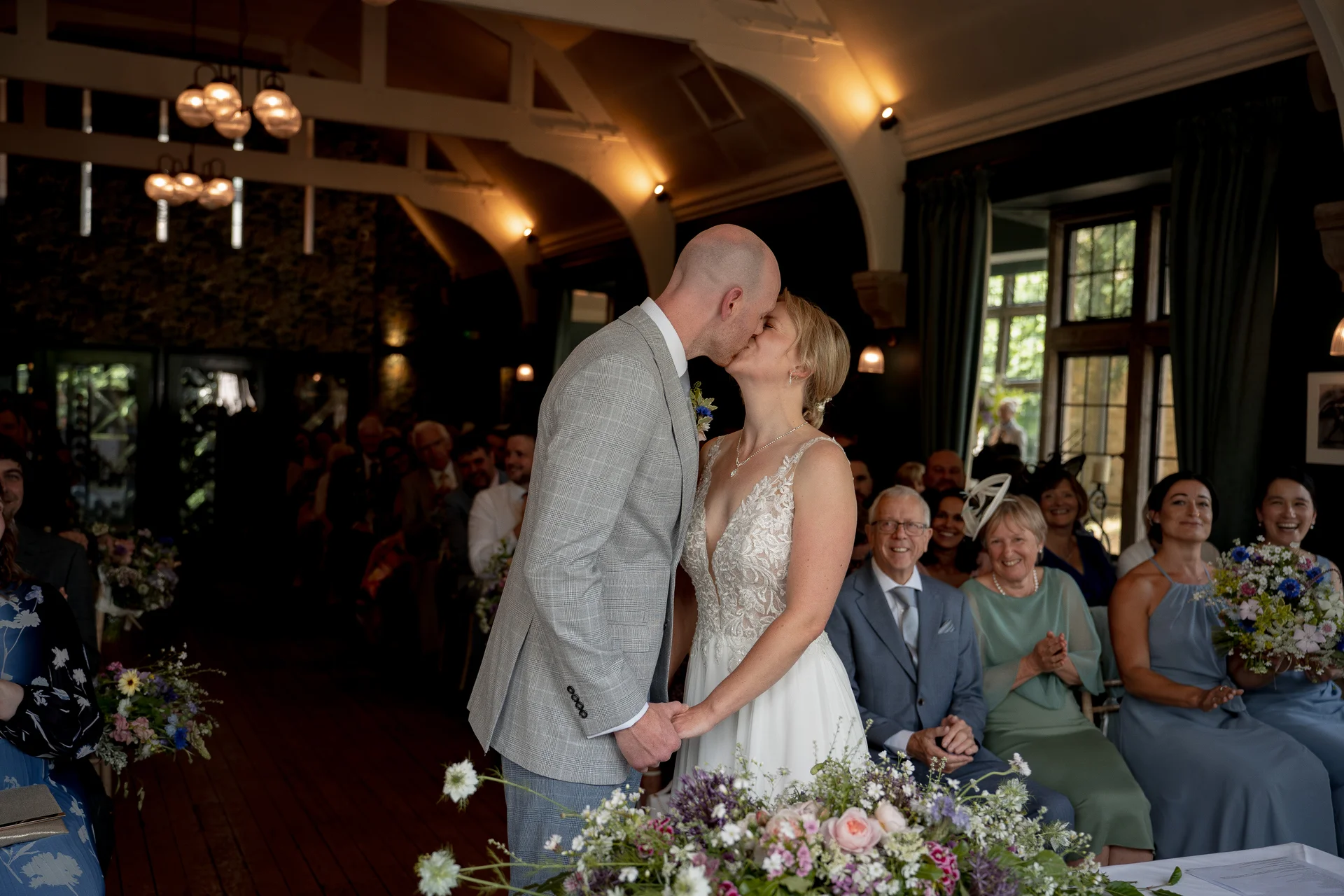 Newlyweds share their first kiss at the front of a rustic hall while seated guests smile and applaud.