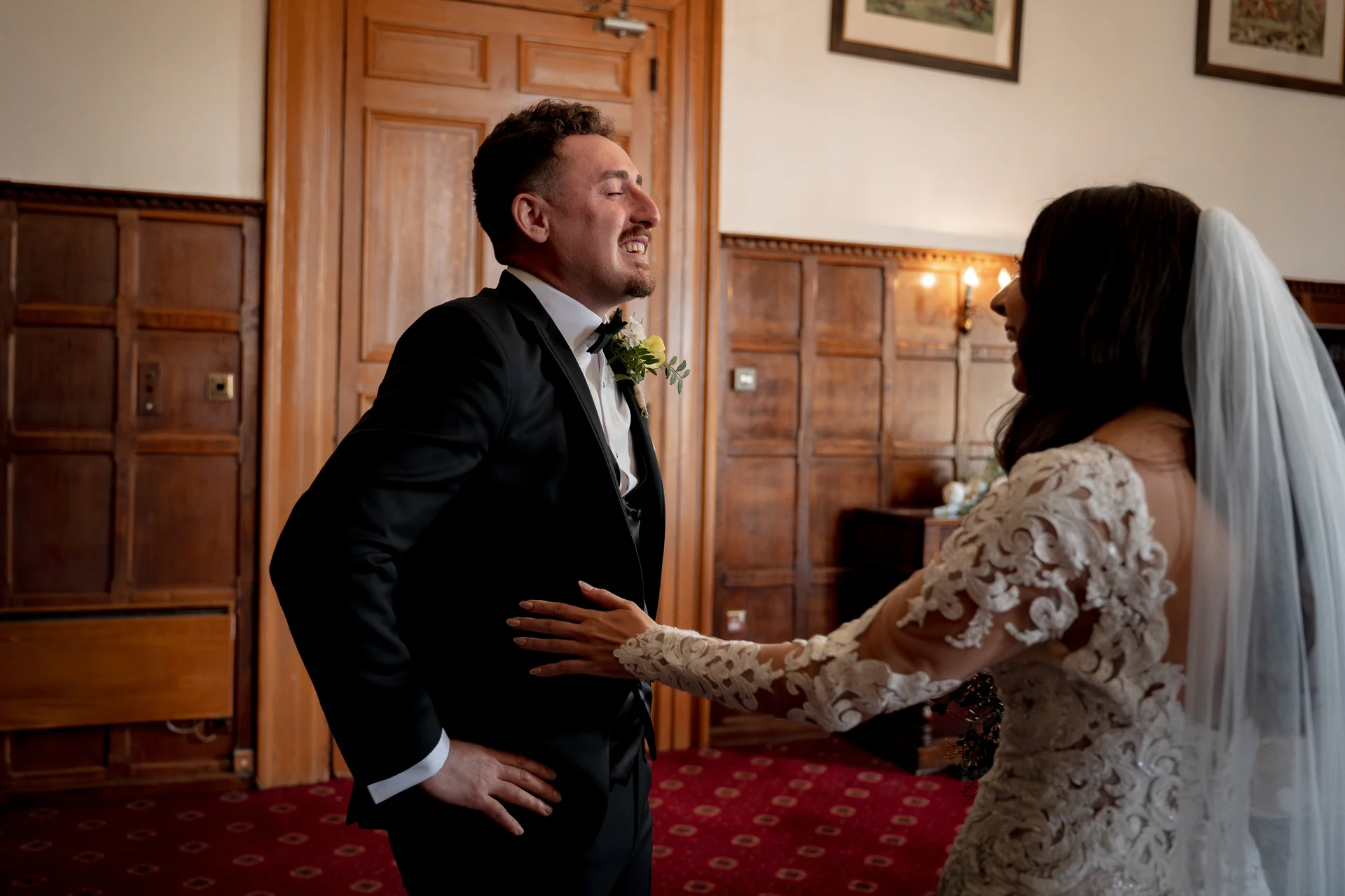 The groom reacts with joyful laughter during a first look moment with his bride inside a wood-panelled room.
