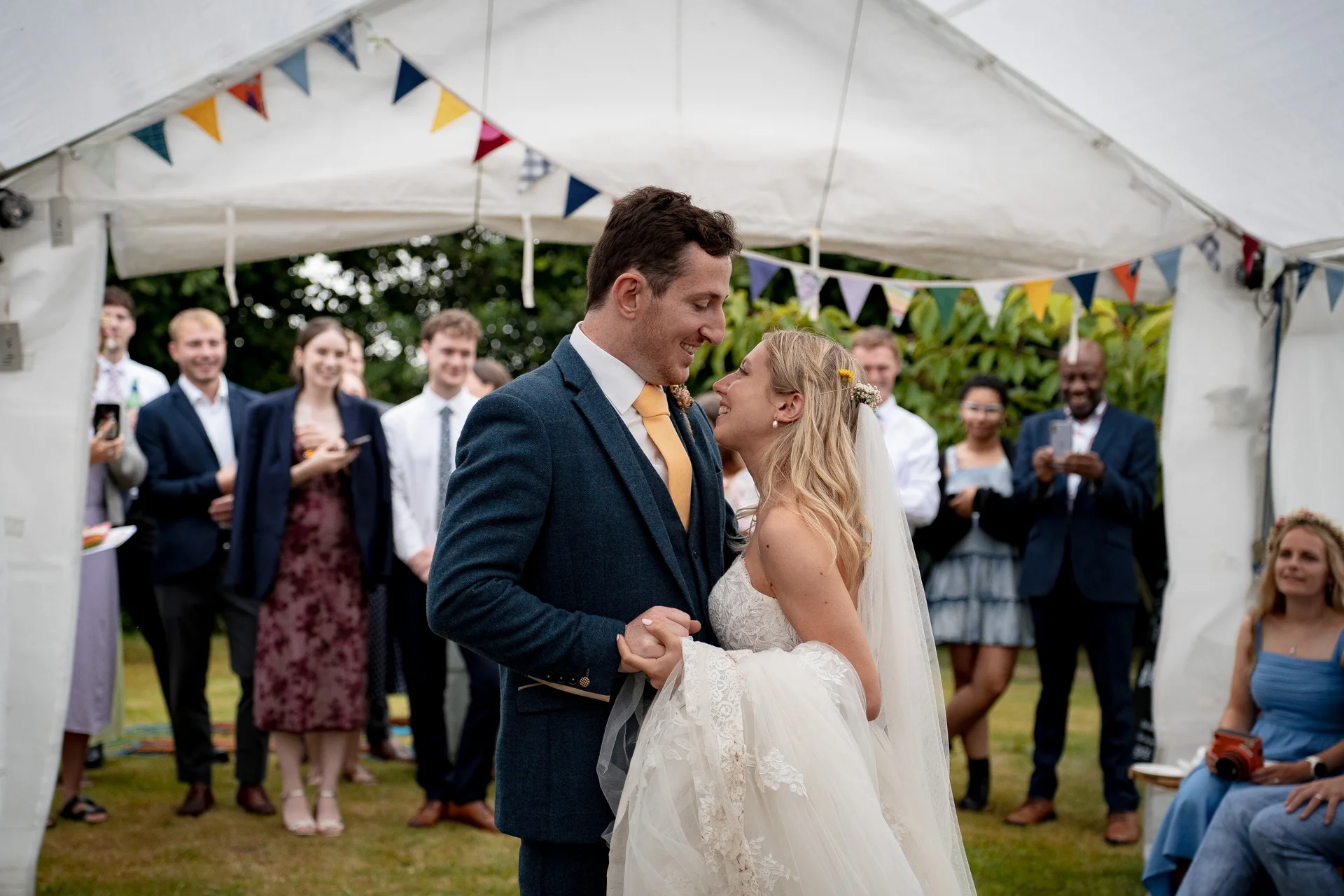 A newlywed couple shares their first dance under a white marquee decorated with colourful bunting, surrounded by smiling wedding guests.