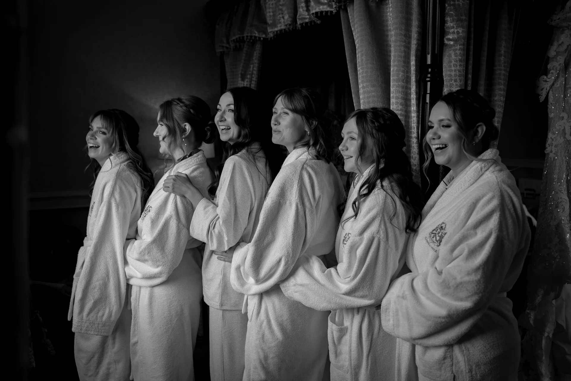 A line of bridesmaids wearing matching white robes stand together smiling and laughing in a room filled with natural window light.