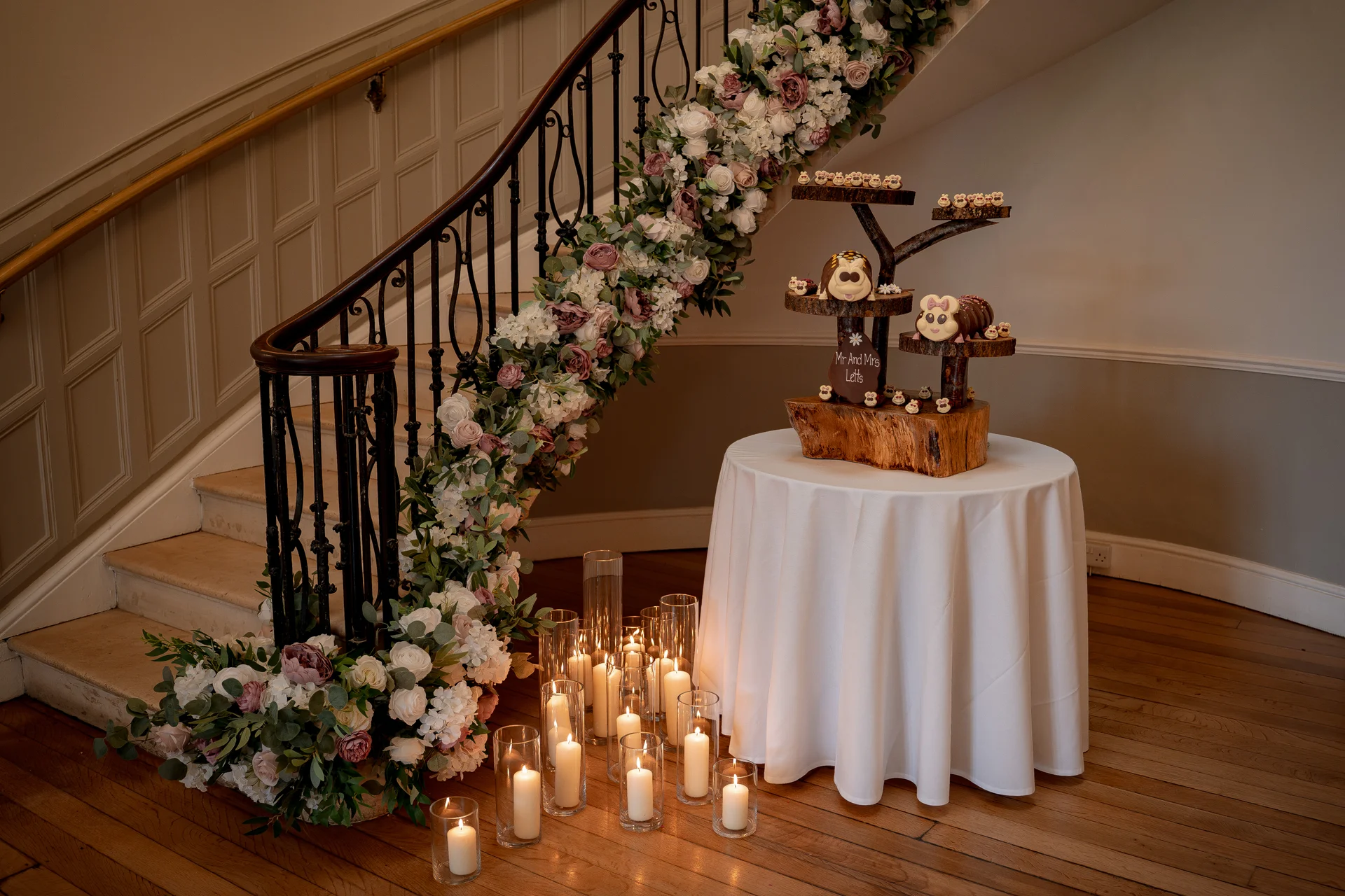 An ornate indoor staircase decorated with a lush floral garland of blush roses and greenery sits beside a round table holding a rustic tree-slice cupcake display surrounded by glowing pillar candles.