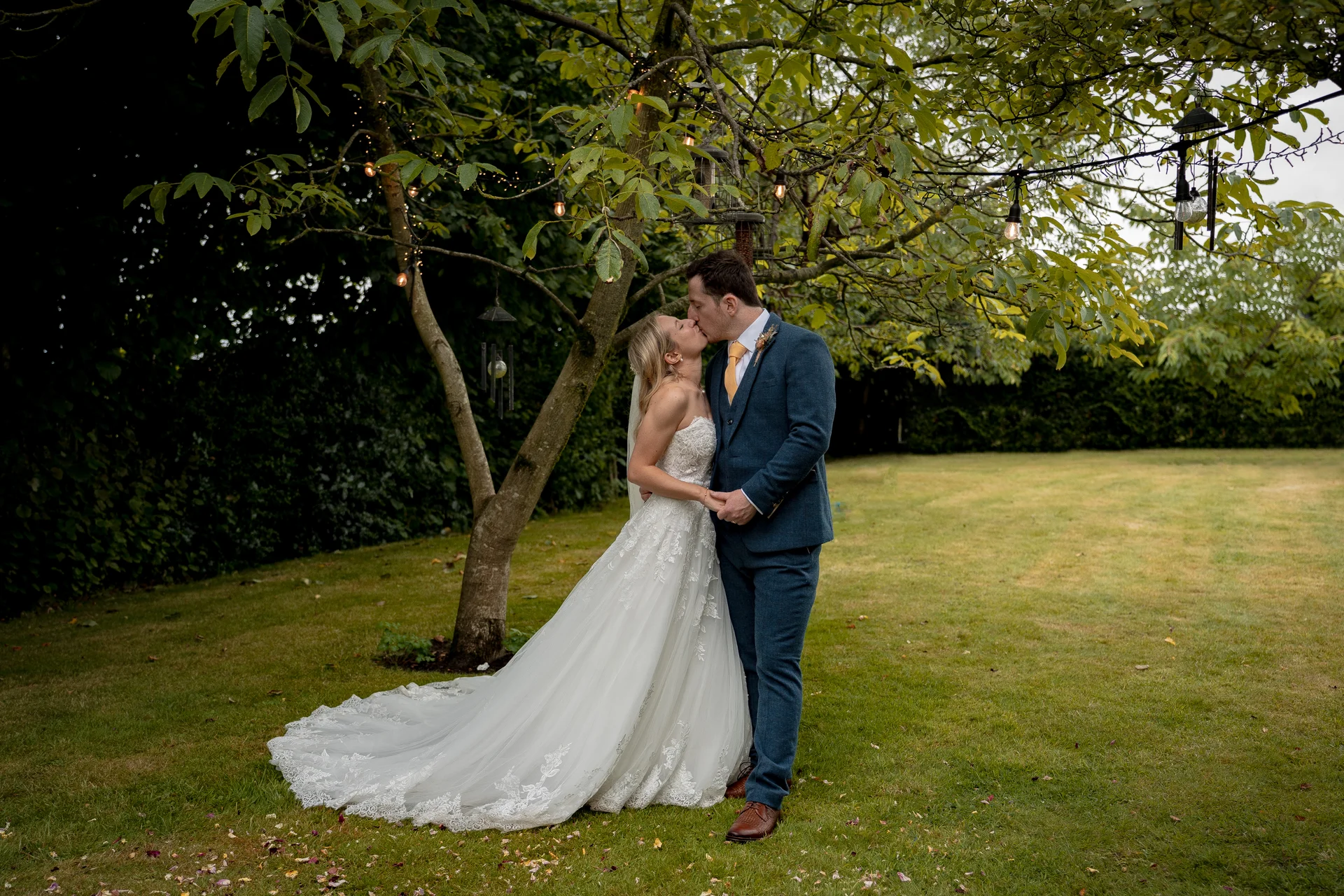 A newlywed couple shares a tender kiss under the branches of a tree adorned with string lights in a lush green garden.