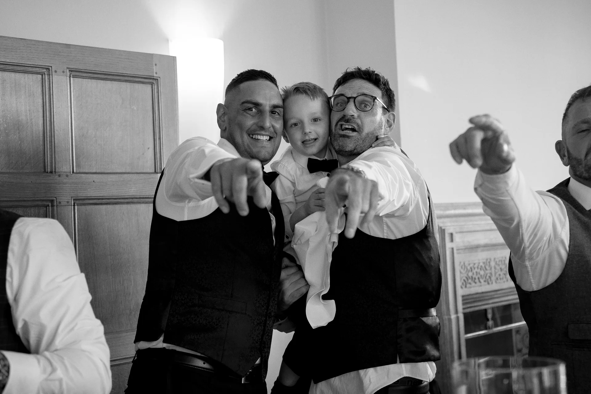 Two smiling groomsmen playfully point towards the camera while holding a young page boy in a matching waistcoat and bow tie during the wedding reception celebrations.