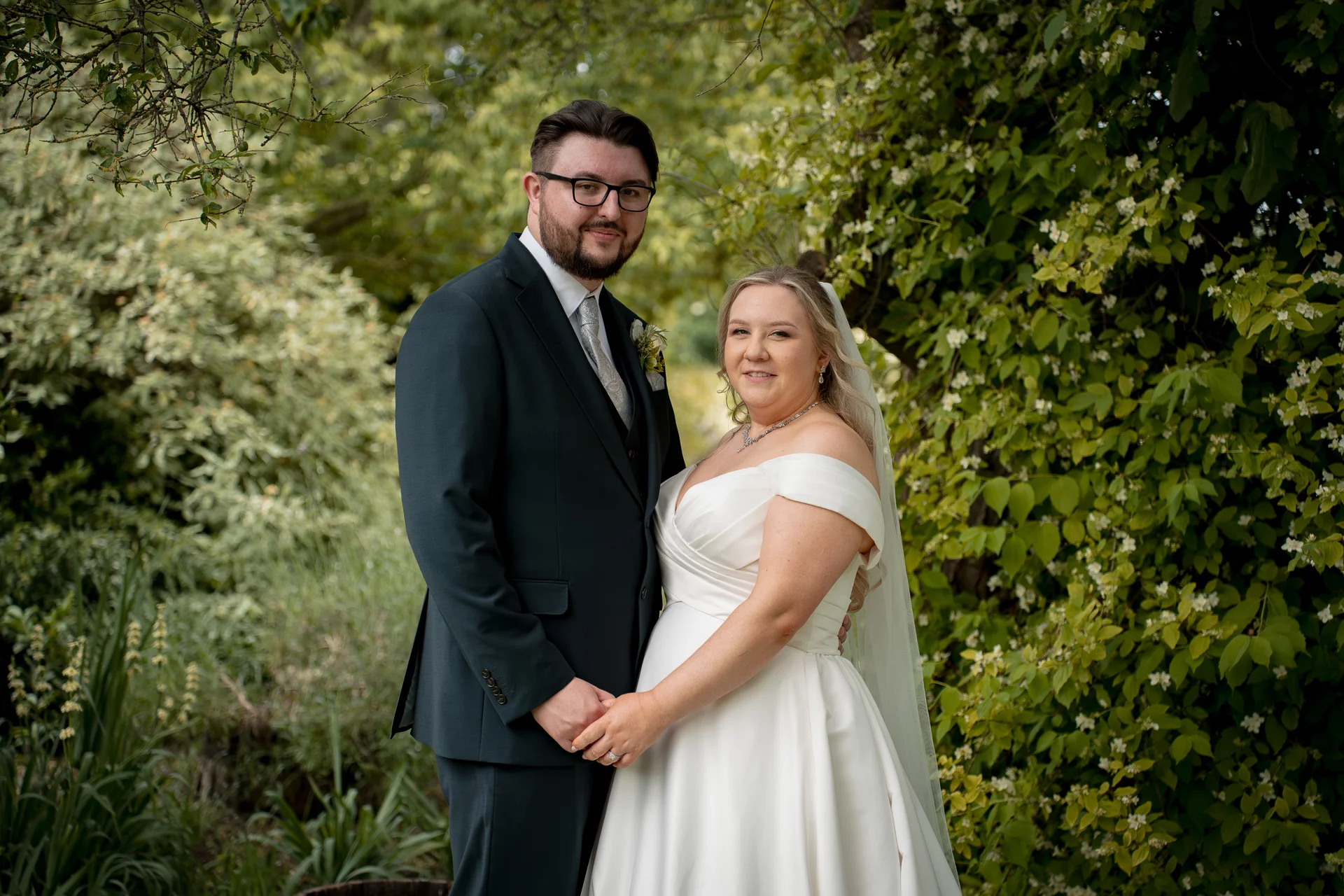 A newlywed couple poses together in a lush green garden, with the bride in an off-the-shoulder satin gown and the groom in a dark suit holding hands.
