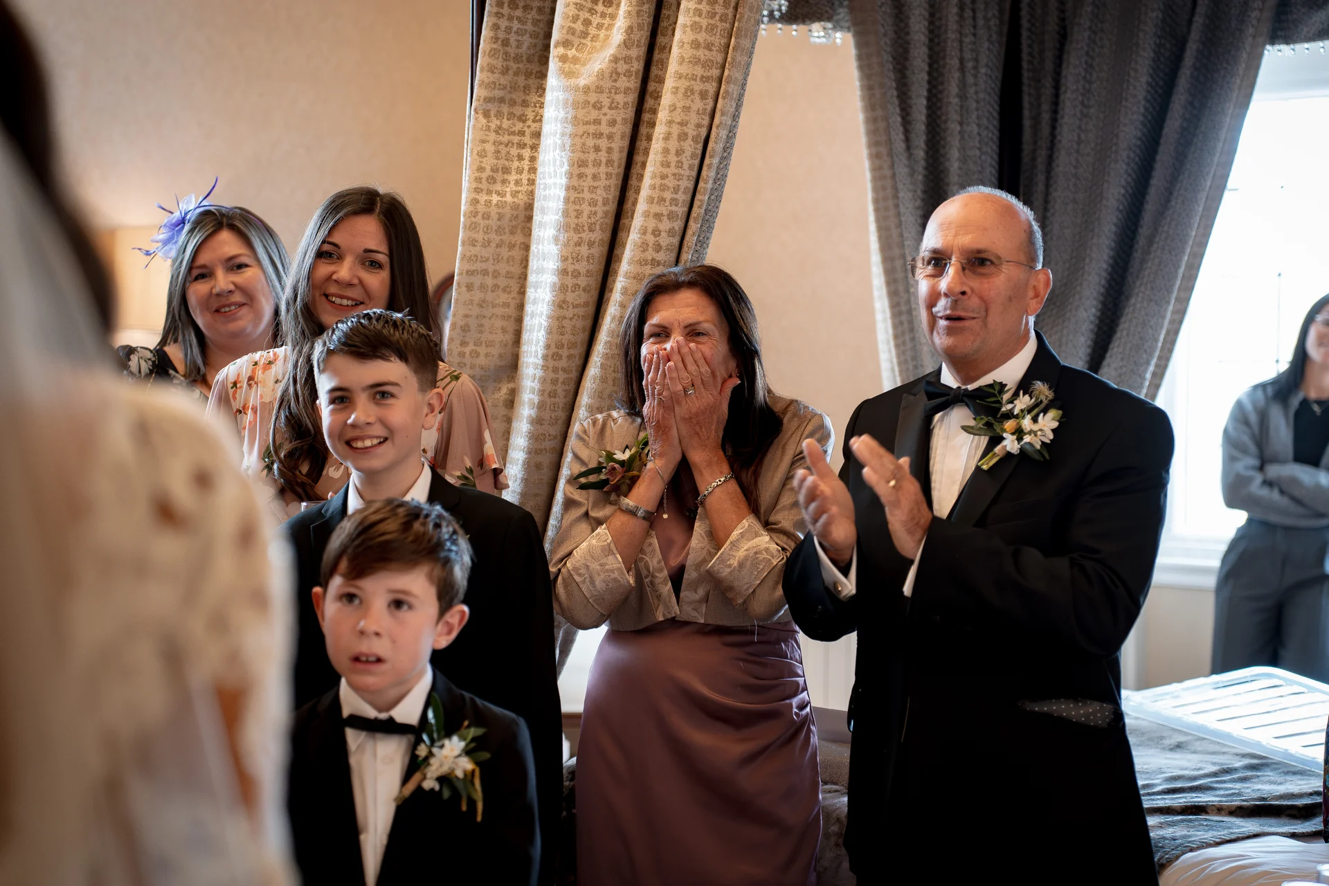 Family members and guests look on with emotional reactions, including a mother covering her face and a father applauding in a tuxedo during a wedding.