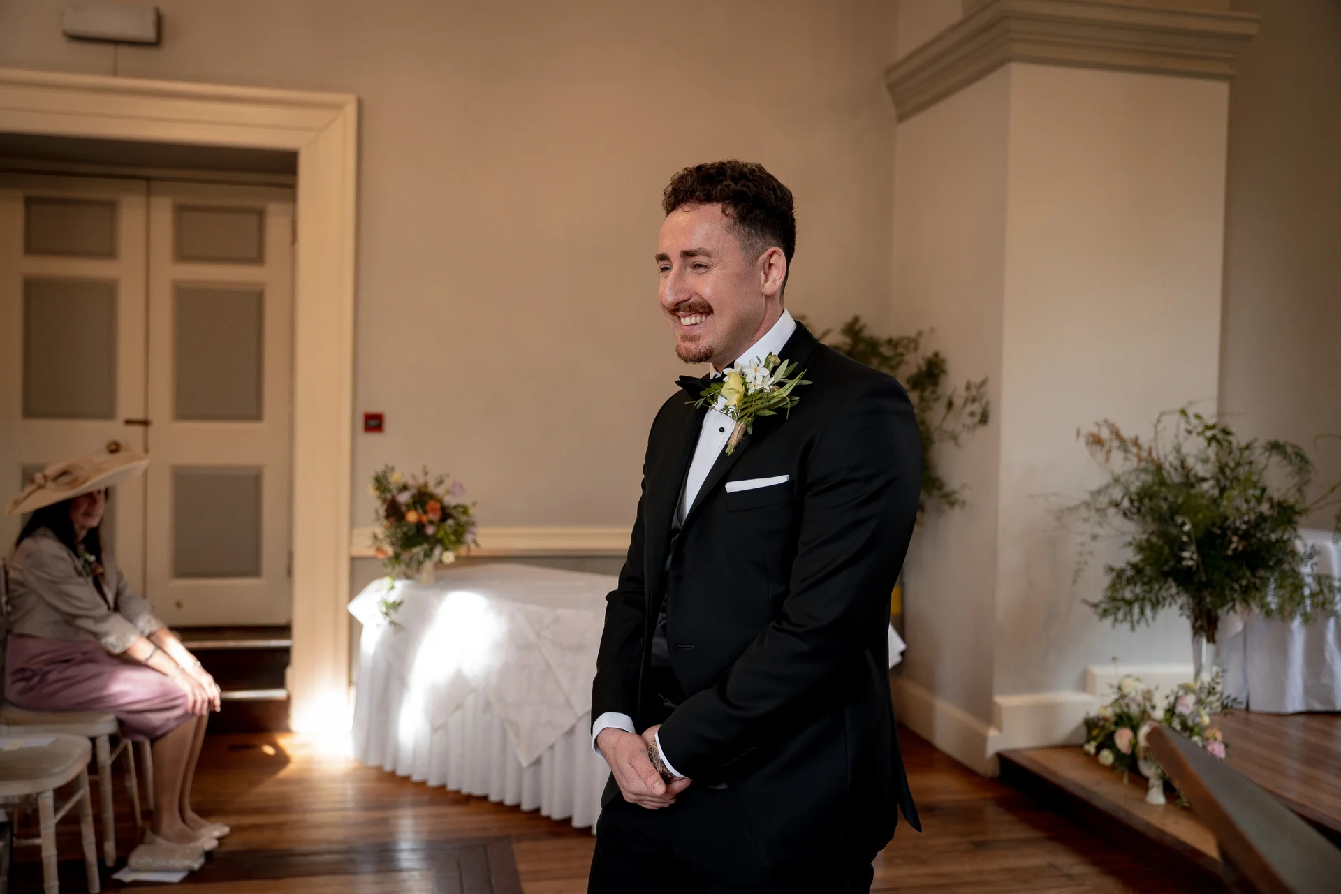 A smiling groom in a classic black tuxedo and bow tie waits at the altar during an indoor ceremony, with a seated guest visible in the background.