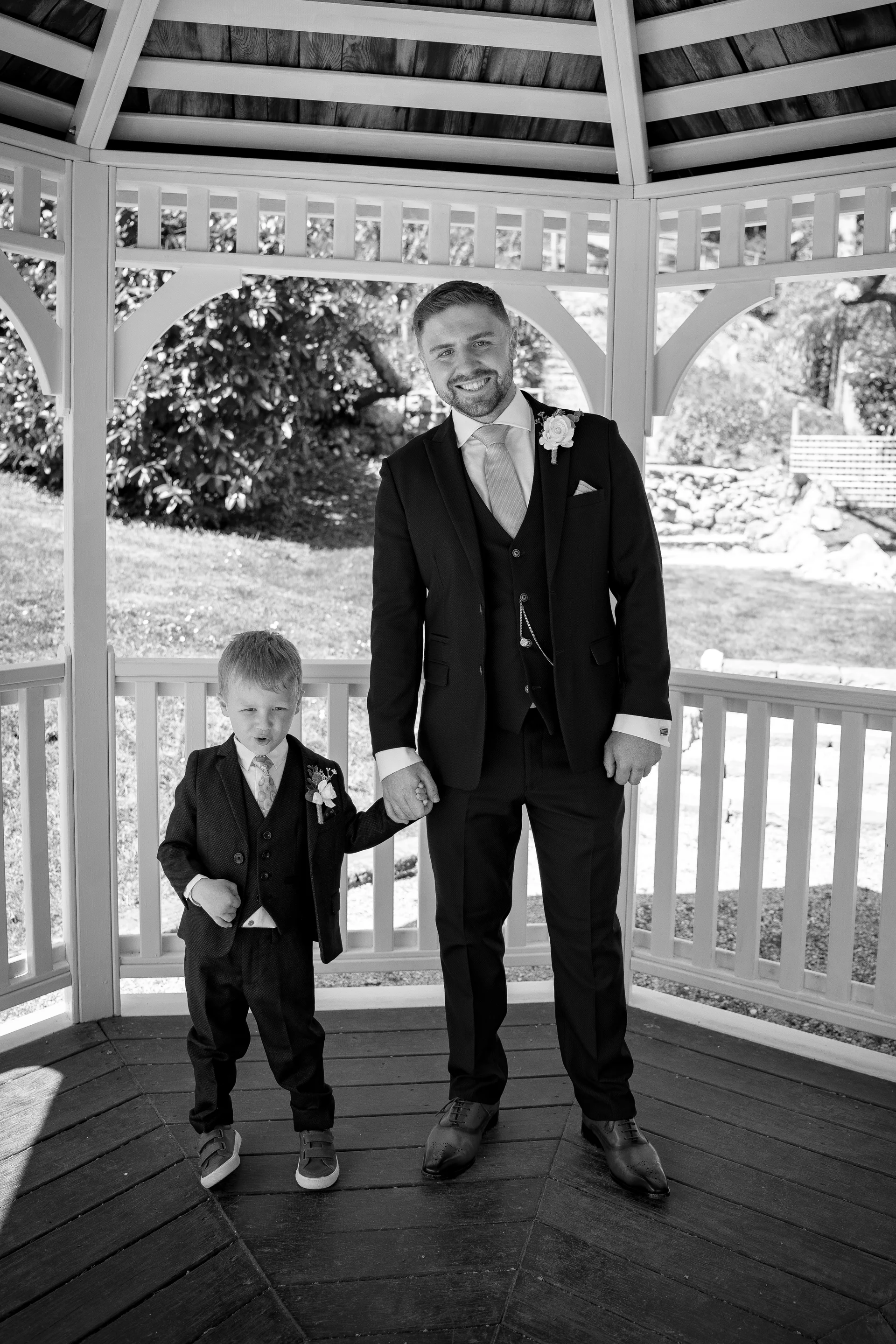 A smiling groom in a three-piece suit stands inside a white wooden gazebo, holding hands with a young page boy who is also dressed in a smart matching suit.