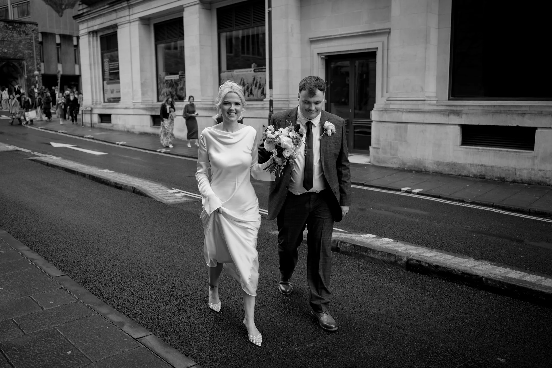 A newlywed couple smiles happily as they walk together along a city street, with the bride holding a bouquet and wearing a sleek satin gown.