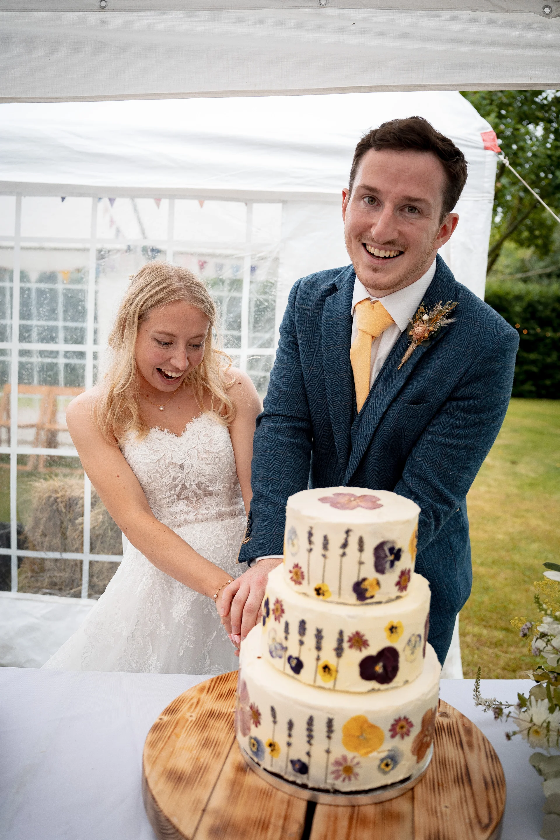 A smiling bride and groom excitedly cut a three-tier pressed flower wedding cake inside a marquee with a wooden stand.