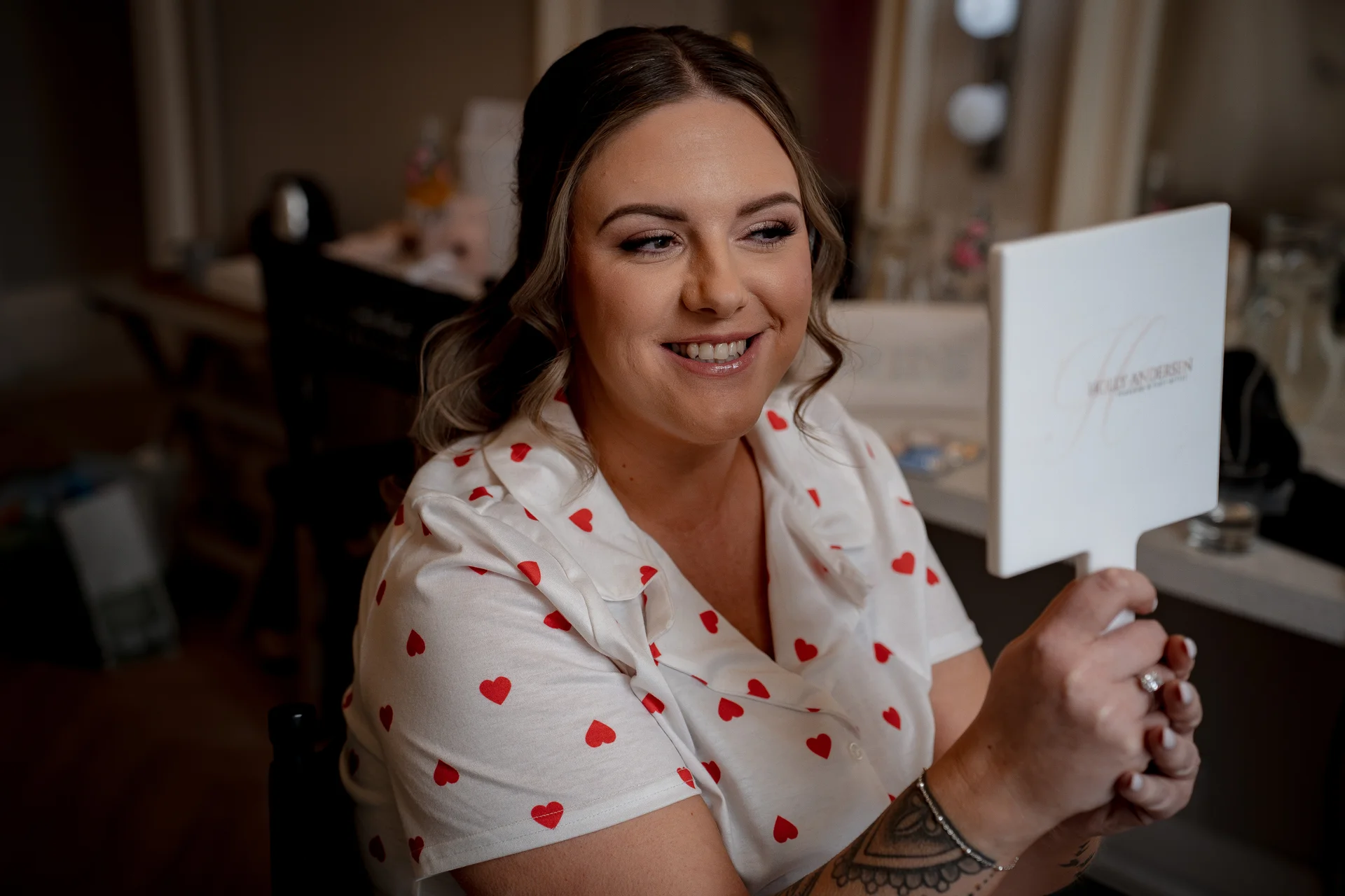 A smiling bride wearing heart-print pyjamas checks her makeup in a handheld mirror during morning bridal preparations.