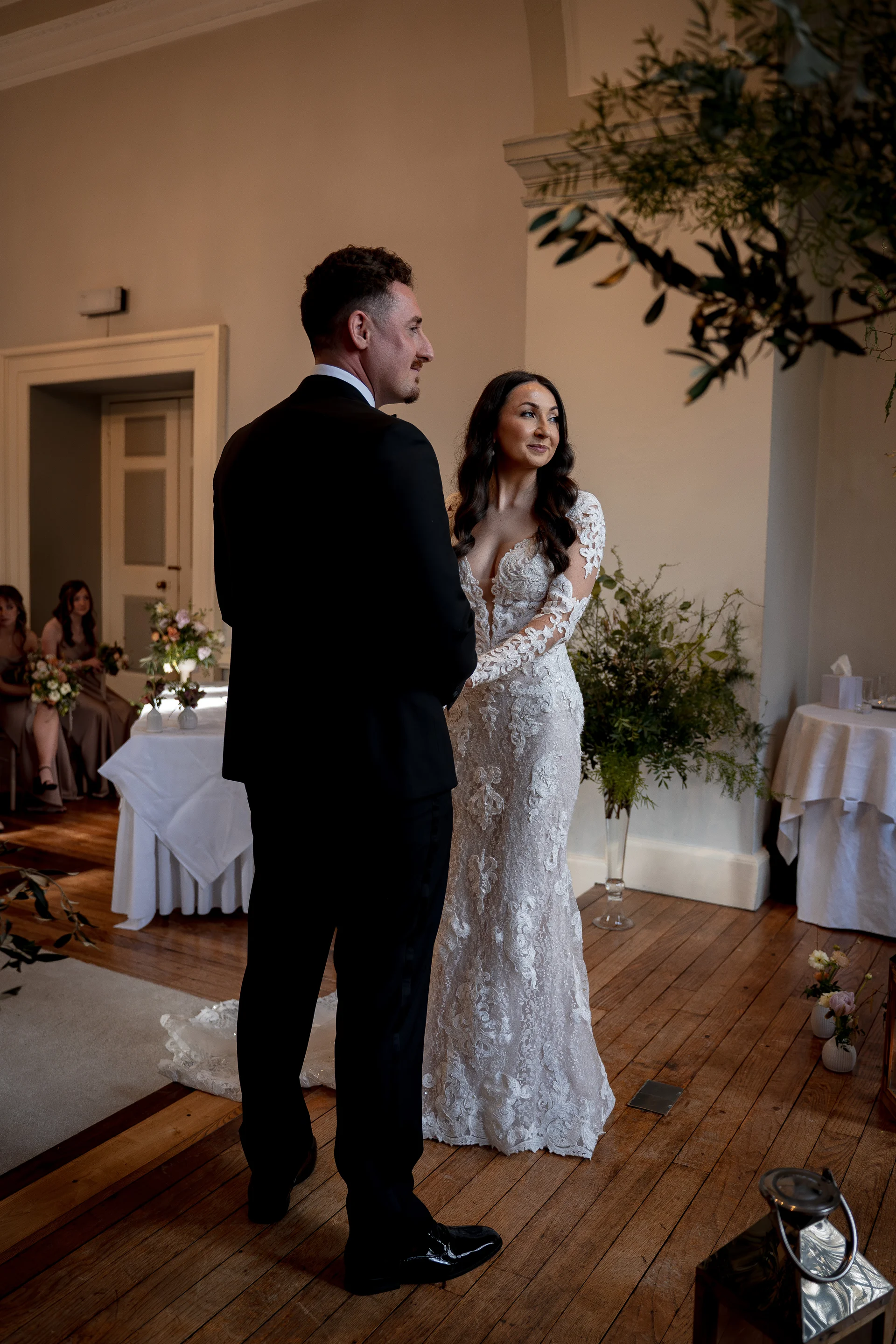 A newlywed couple stands together indoors during their wedding ceremony, with the groom in a classic black tuxedo and the bride in a form-fitting lace long-sleeve gown.