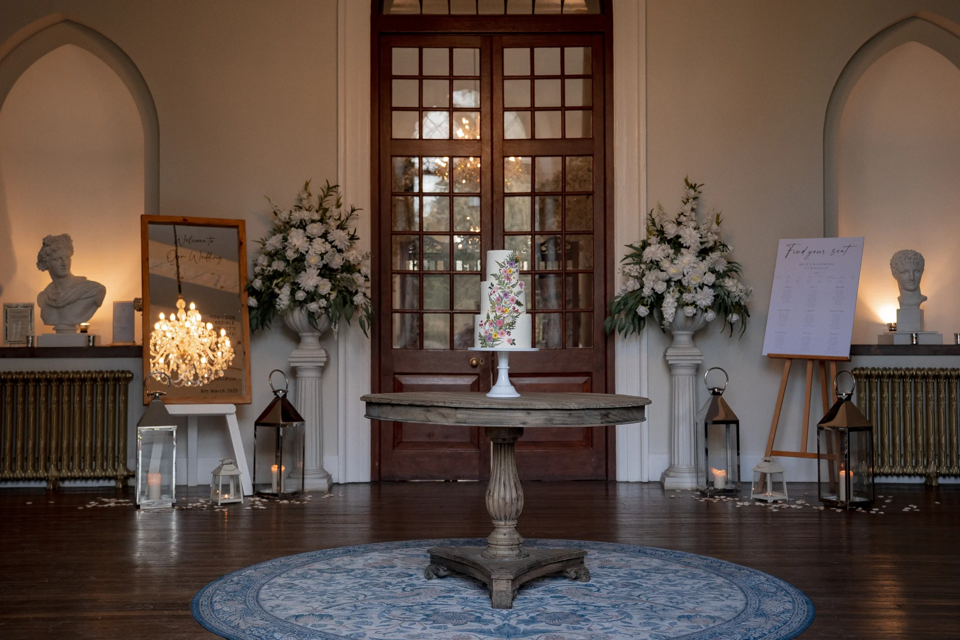 A three-tier hand-painted wedding cake sits centrally on a wooden pedestal table inside a grand manor house, flanked by floral urns and welcoming signage.