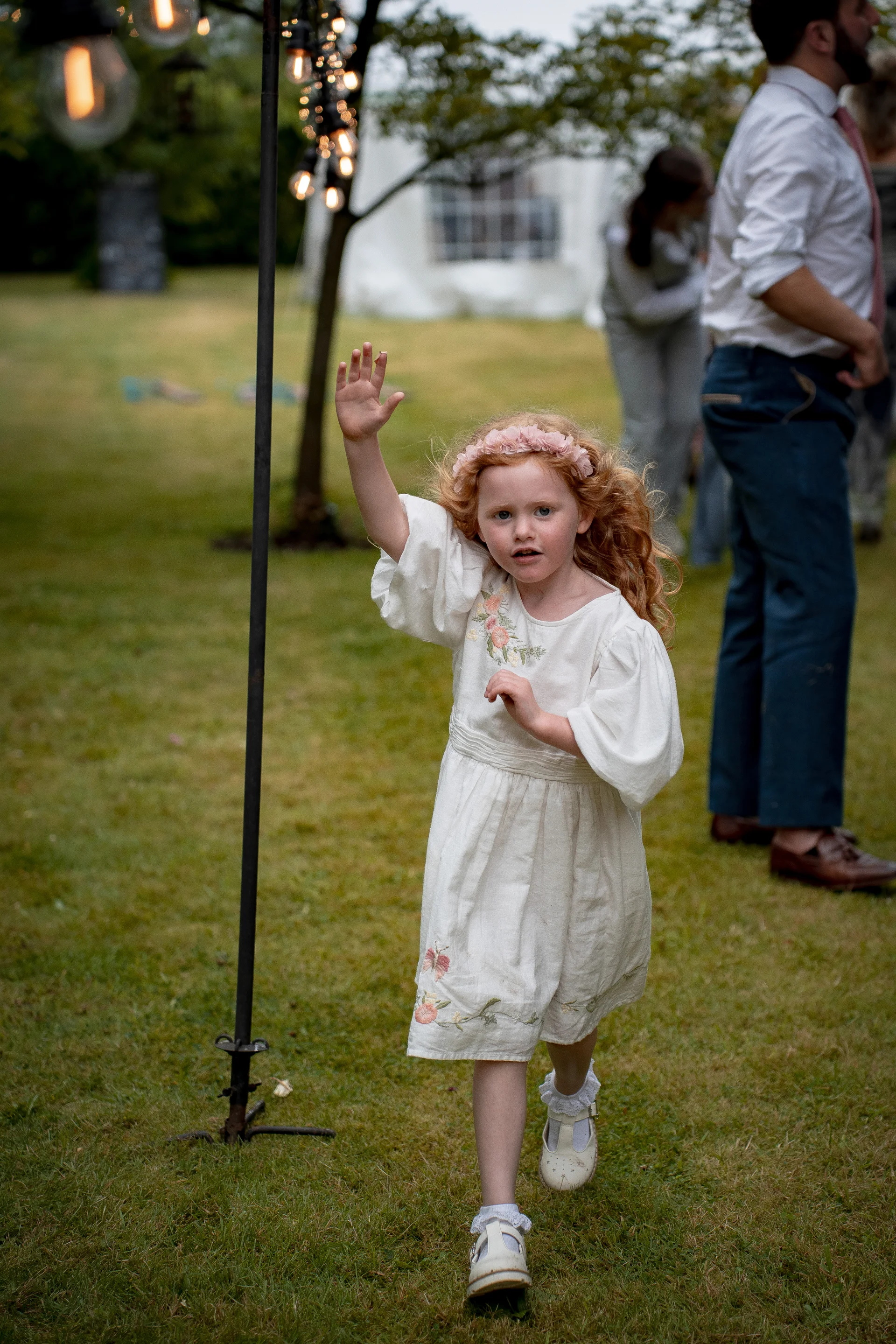 A young flower girl with red hair and a floral crown waves happily while walking across a grassy lawn during an outdoor wedding reception.