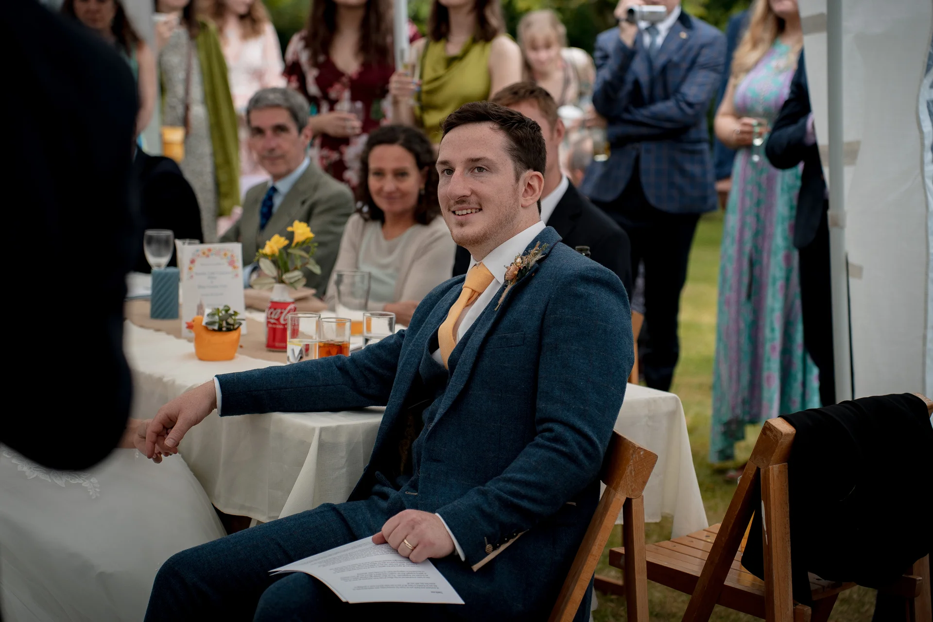 A smiling groom in a blue tweed suit with a yellow tie holds a speech at a wedding breakfast in a marquee.