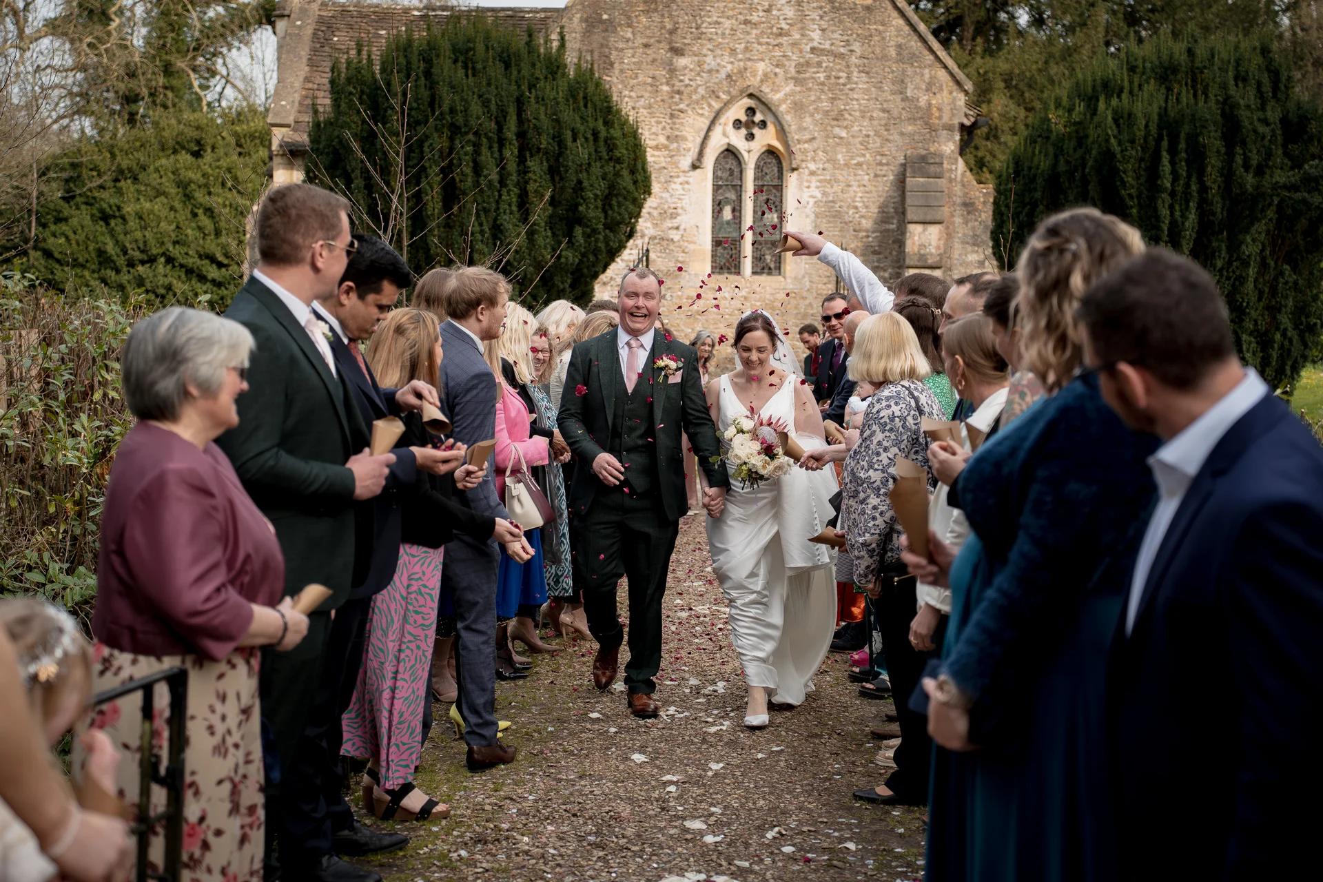 A newlywed couple laughing joyfully as wedding guests throw confetti over them on a gravel path outside a historic stone church.