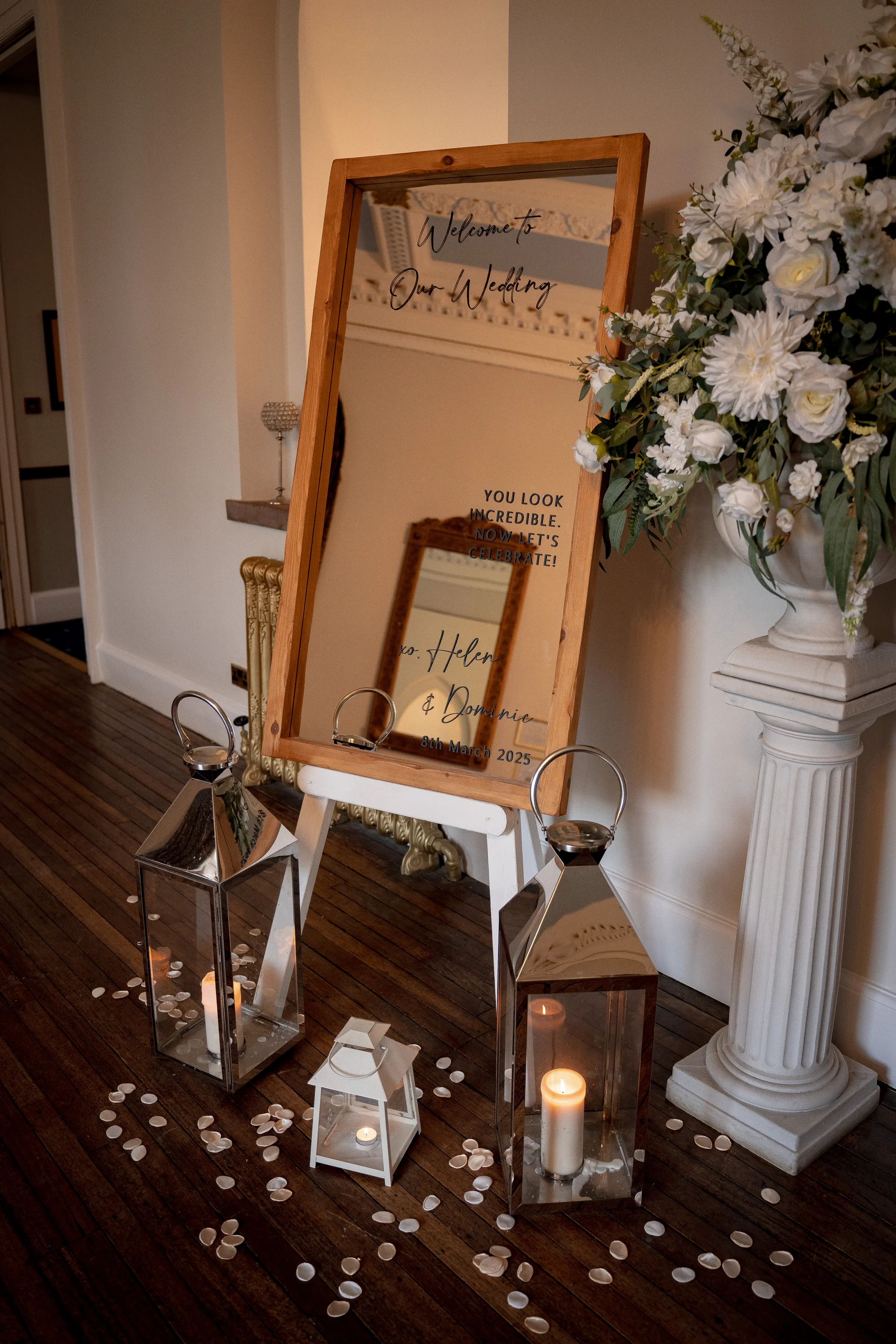 An elegant wedding welcome sign on a mirror displayed on an easel, surrounded by lit lanterns and white rose floral arrangements.