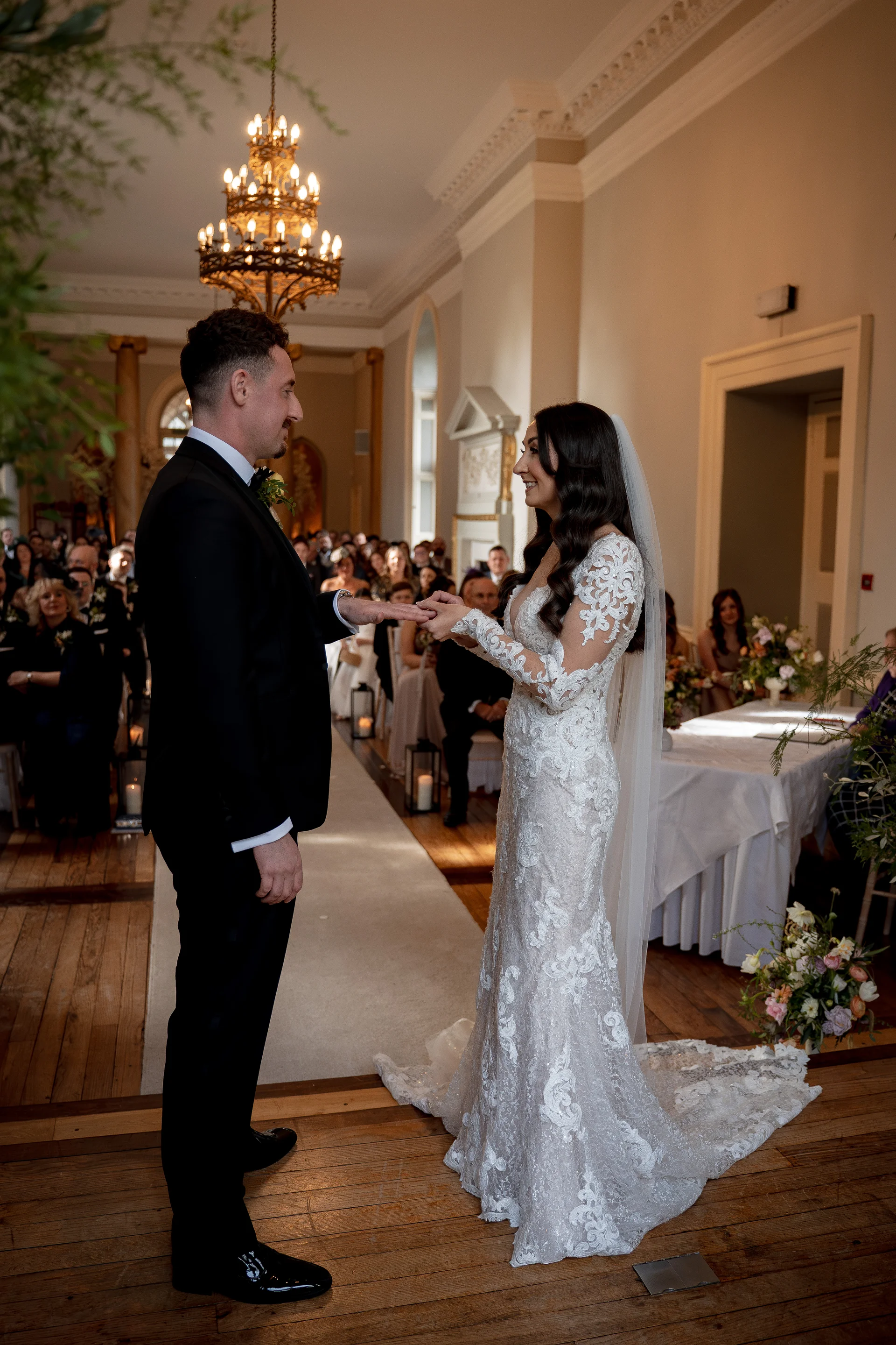 A bride and groom exchange rings during their indoor wedding ceremony in a grand room featuring wooden floors, a large chandelier, and seated guests watching.
