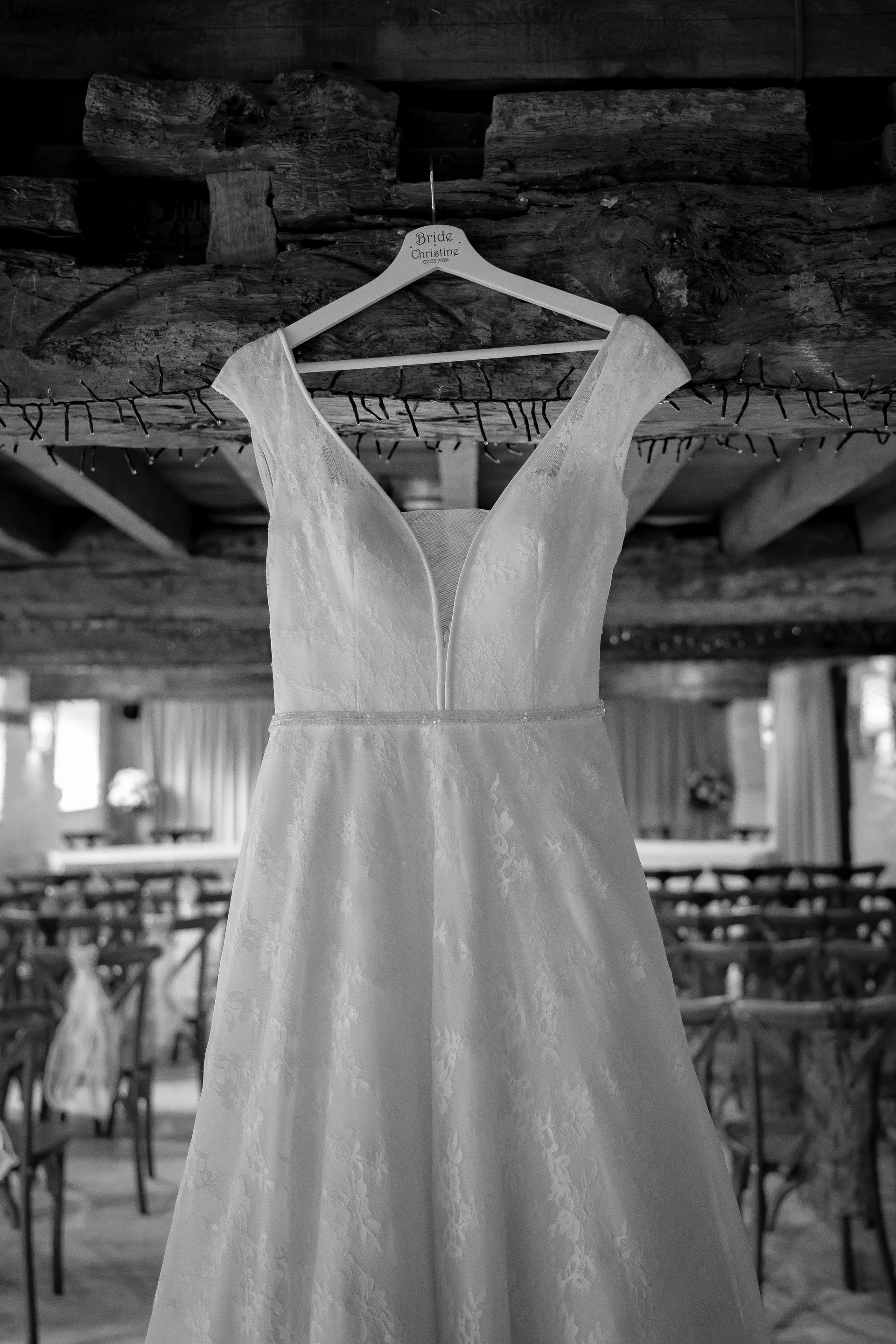 A lace wedding dress with a V-neckline hangs from a personalised wooden hanger against a rustic barn beam backdrop during morning preparations.