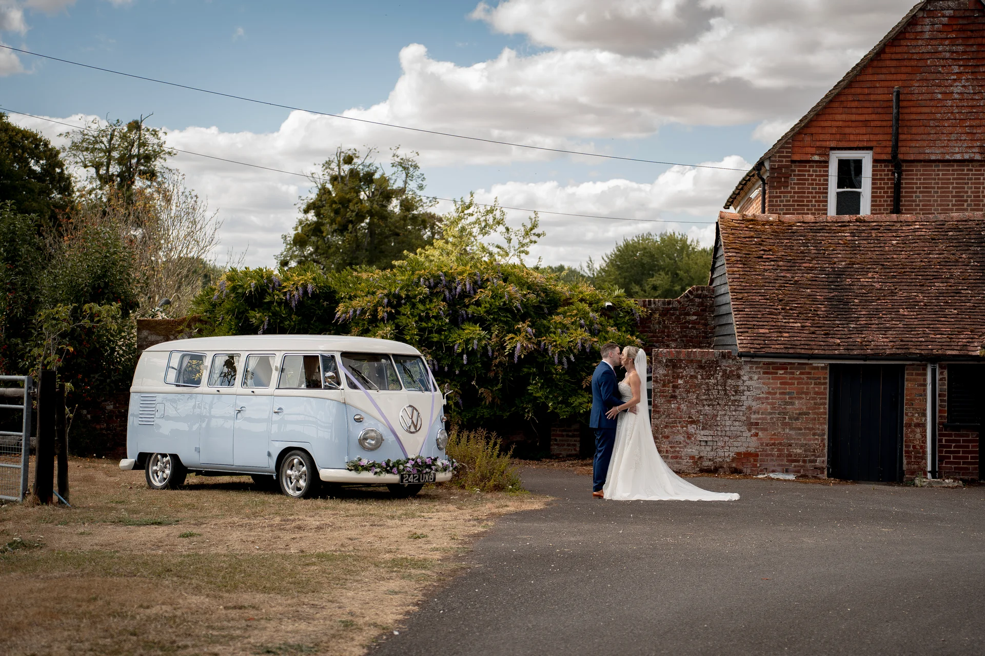 A bride and groom share a tender kiss outside a rustic brick barn next to a vintage powder blue Volkswagen campervan on their wedding day.