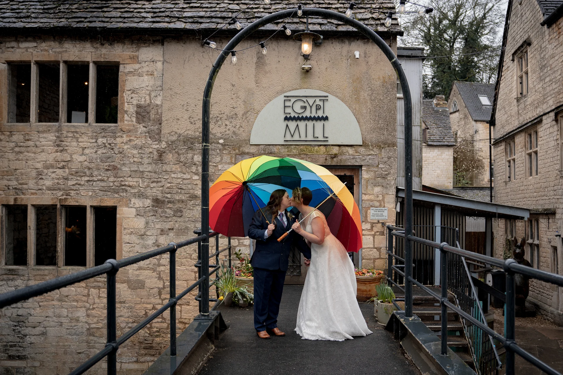 A same sex couple shares a kiss beneath a vibrant rainbow umbrella on a stone bridge outside a historic mill building on a rainy wedding day.