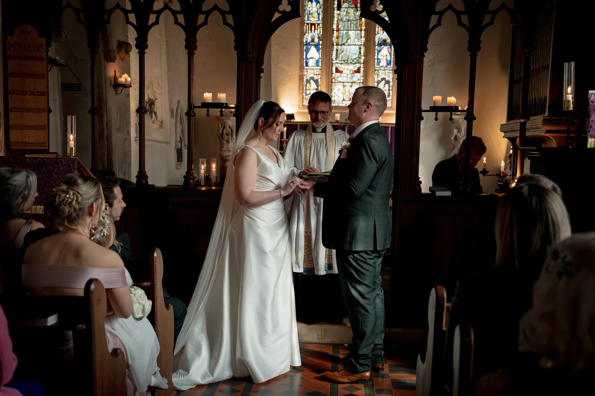 A bride in a satin v-neck gown and a groom in a dark suit exchange rings during a church ceremony illuminated by candlelight and stained glass.