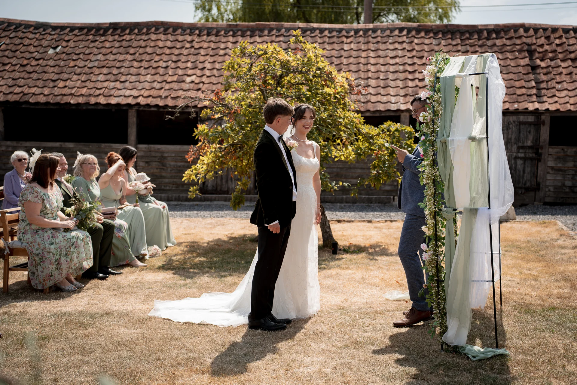 A smiling bride and groom stand together during an outdoor ceremony in front of a rustic barn venue with a floral and fabric arch.