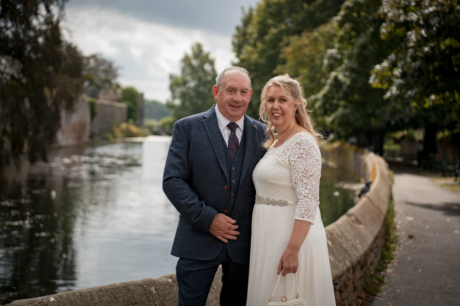 A mature bride and groom pose smiling together on a stone bridge beside a calm river with trees in the background.