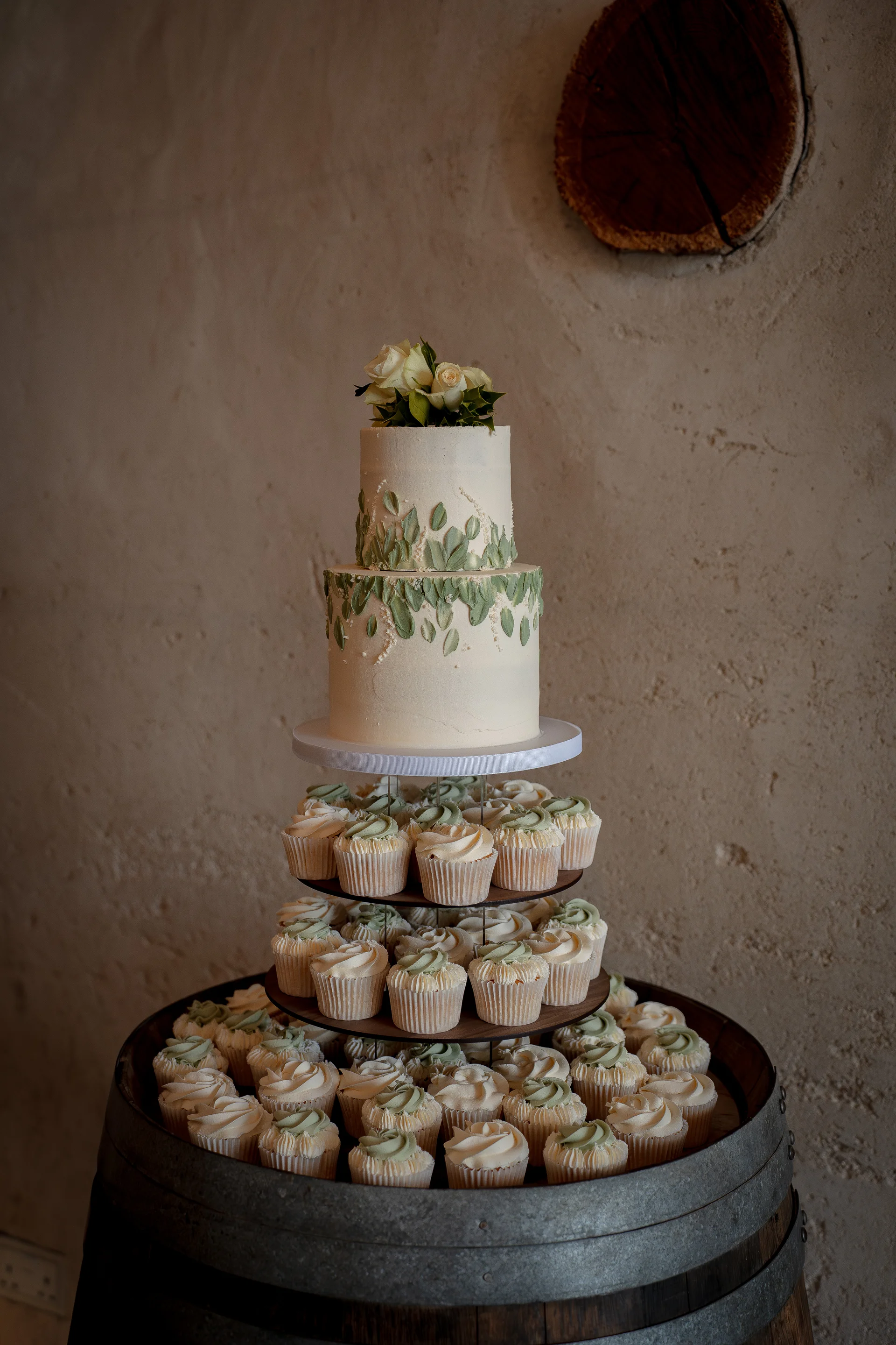 A stunning two-tiered wedding cake with green leaf painted details and fresh roses sits atop a multi-tiered cupcake tower resting on a rustic wine barrel against a textured stone wall.