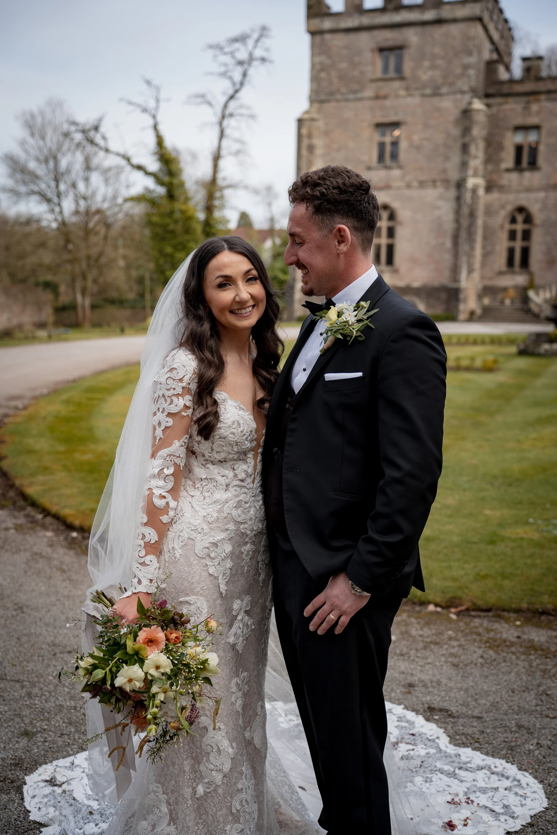 A smiling bride and groom stand outdoors on a pathway with a historic stone castle in the background, the bride holding a hand-tied bouquet and wearing a lace long-sleeve gown.