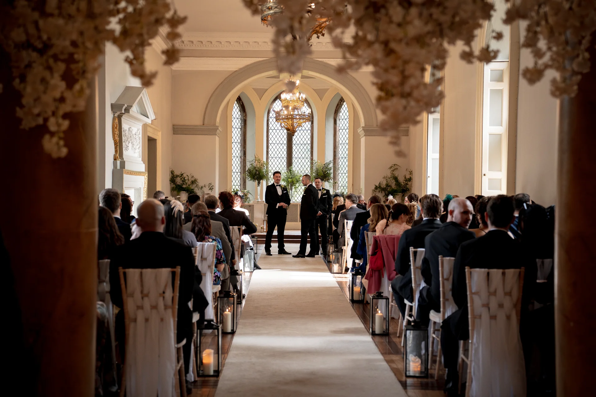 Two grooms stand at the altar in a grand, sunlit historic hall lined with seated guests, framed by hanging floral installations and aisle lanterns.