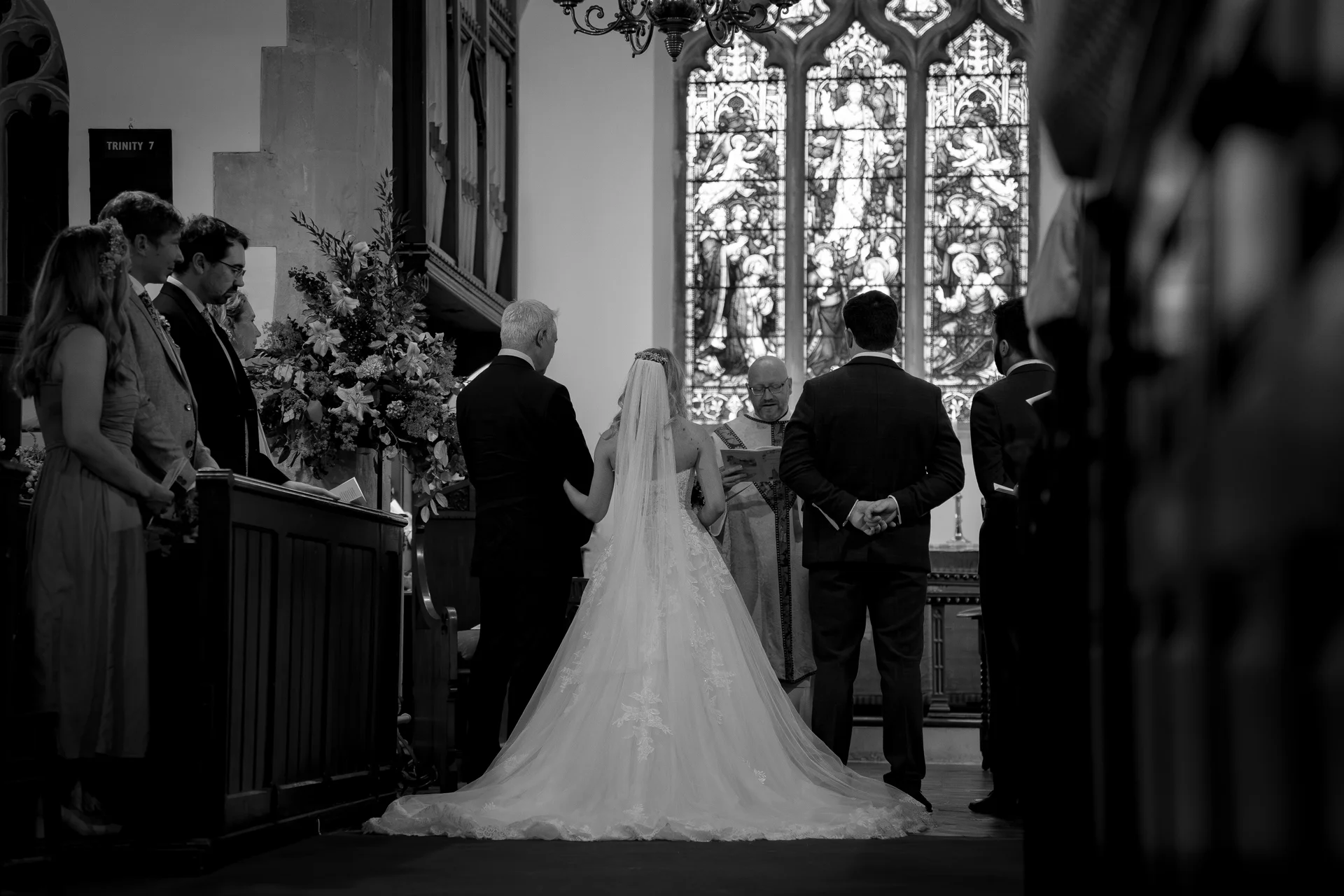 A bride with a dramatic train and veil stands at the altar during a traditional church wedding ceremony beneath stained glass windows.