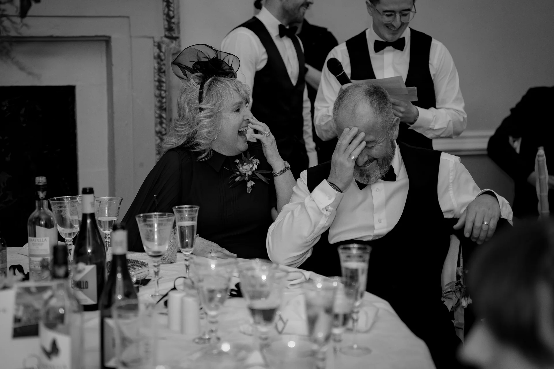 A guest wearing a fascinator laughs hysterically while covering her mouth at the wedding reception table, alongside a man holding his head in amusement as speeches take place.
