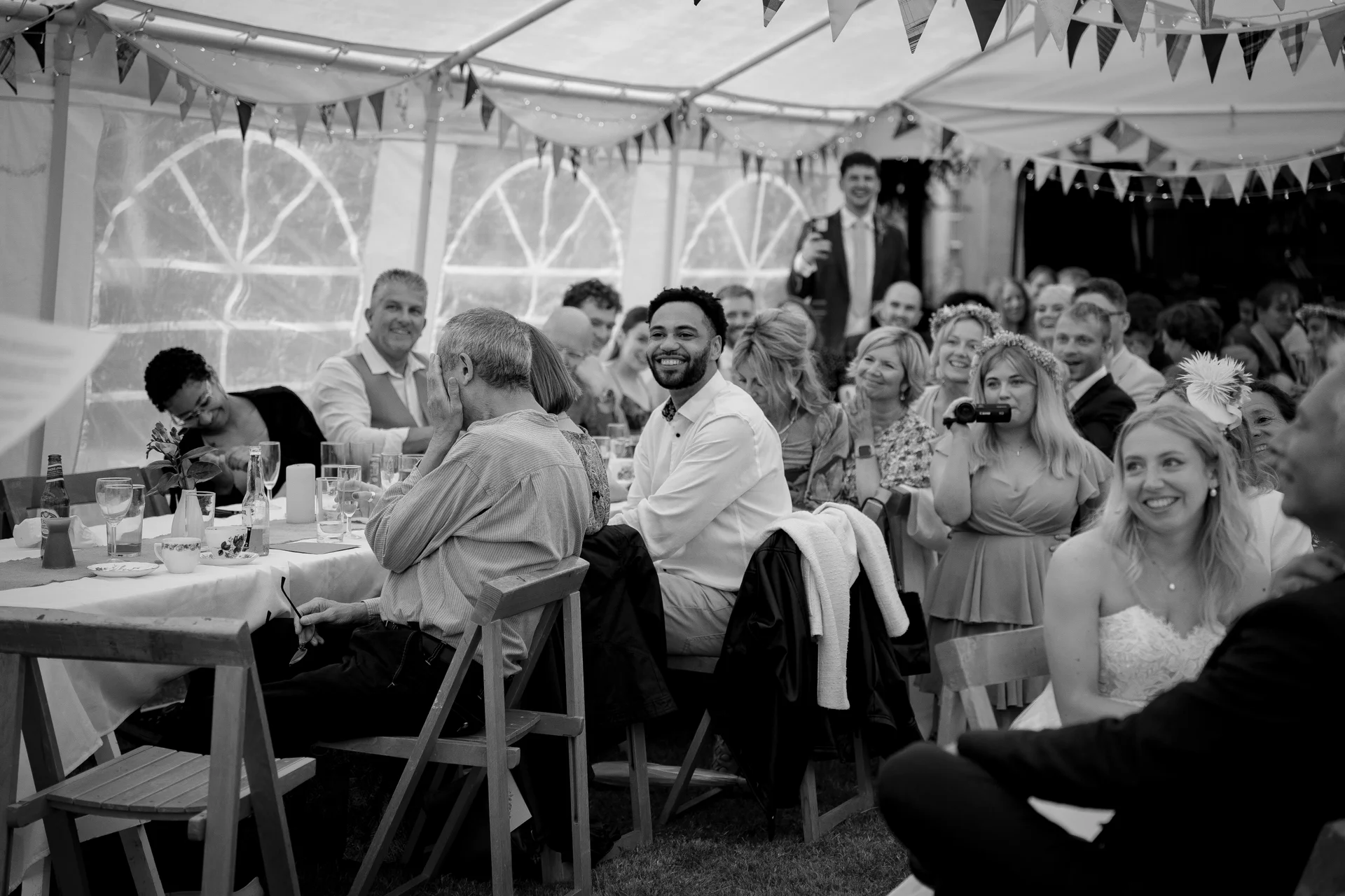 Guests seated at long tables inside a marquee laugh and enjoy themselves during the wedding speeches.