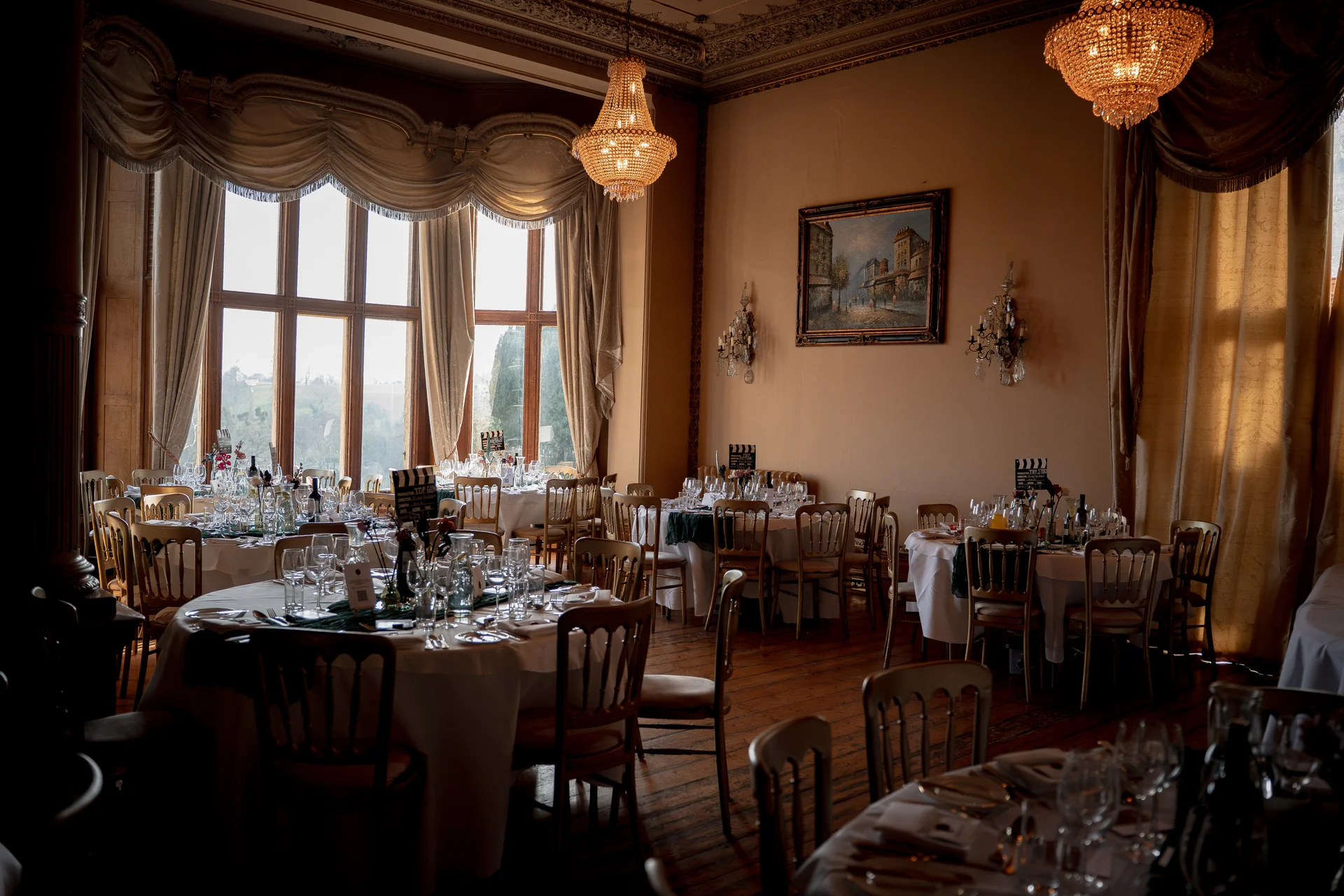 An elegant wedding reception room featuring round dining tables set with crisp white linens and crystal glassware beneath ornate chandeliers and large arched windows.