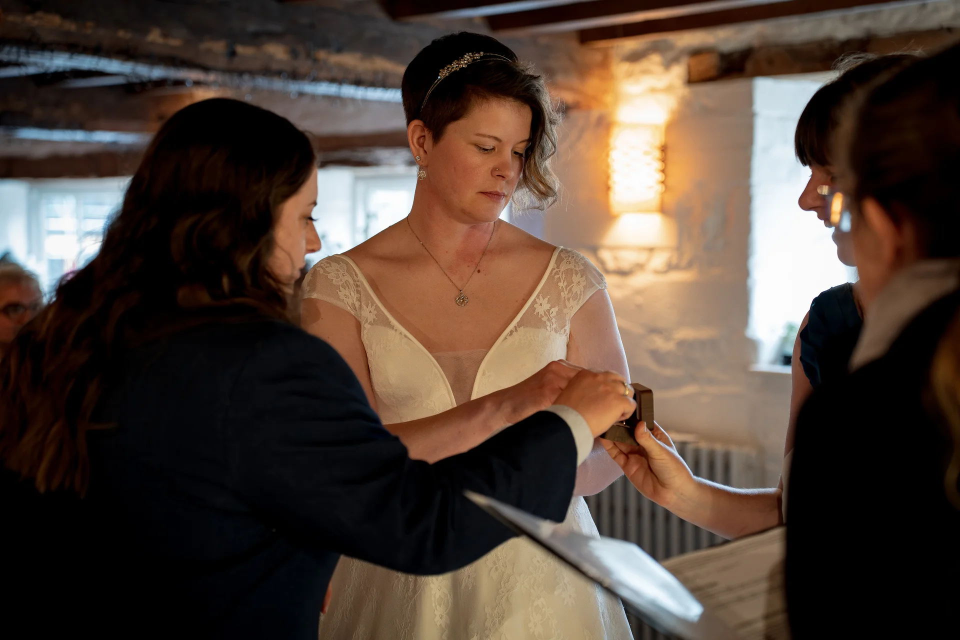 A short-haired bride looks down attentively during the ring exchange ceremony inside a rustic stone barn with exposed wooden rafters.