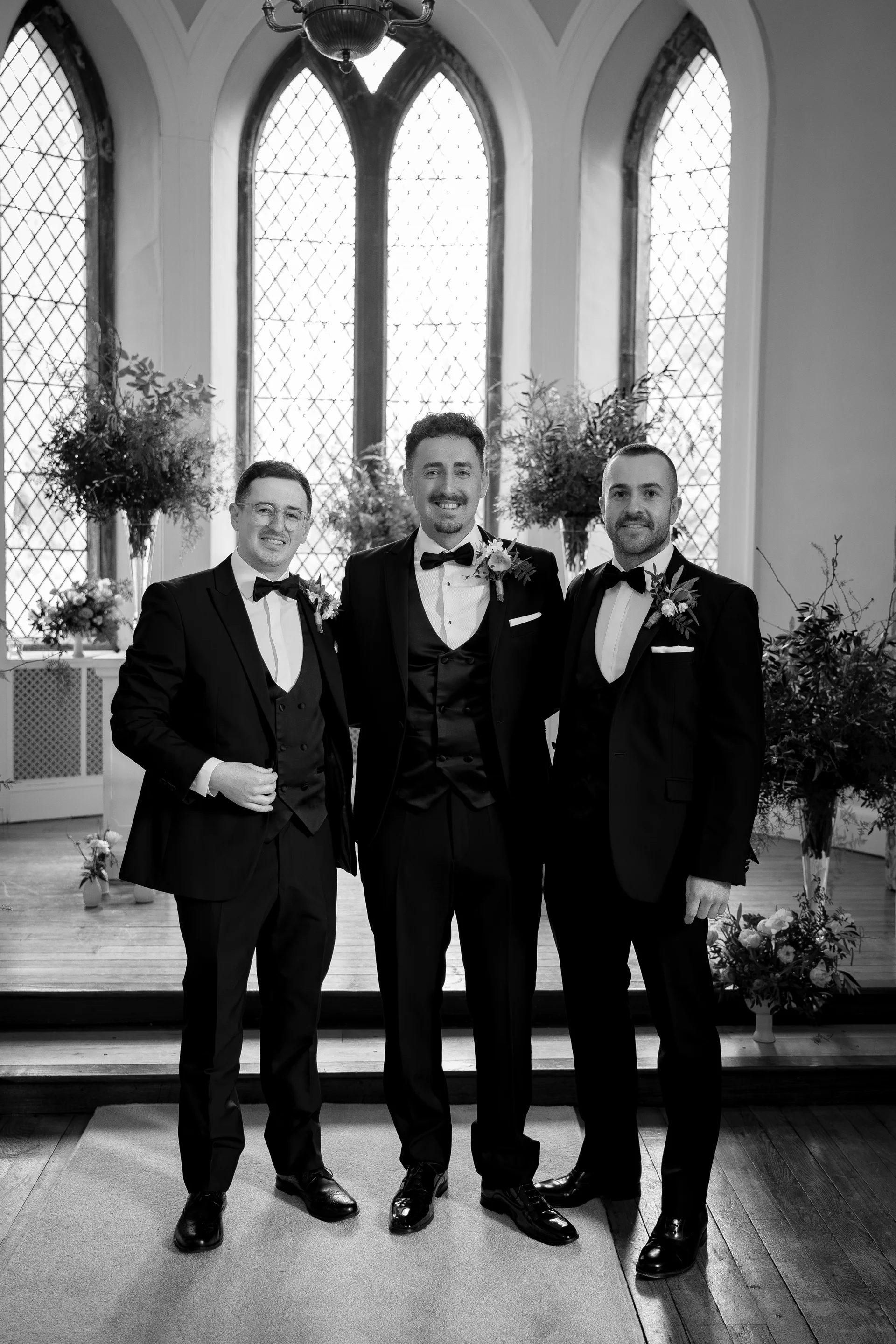Three smiling groomsmen in classic black tuxedos and bow ties pose inside a bright church with tall arched stained glass windows.