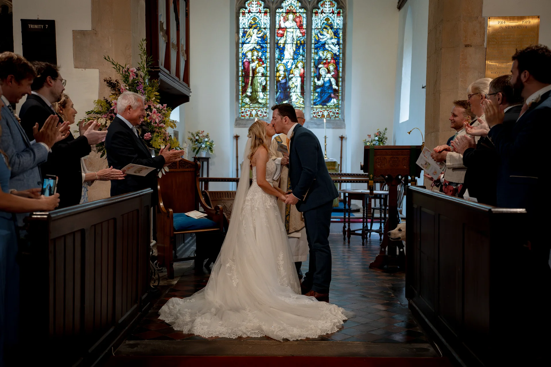 The newly married couple share their first kiss at the altar of a traditional church, surrounded by cheering guests and stained glass windows.