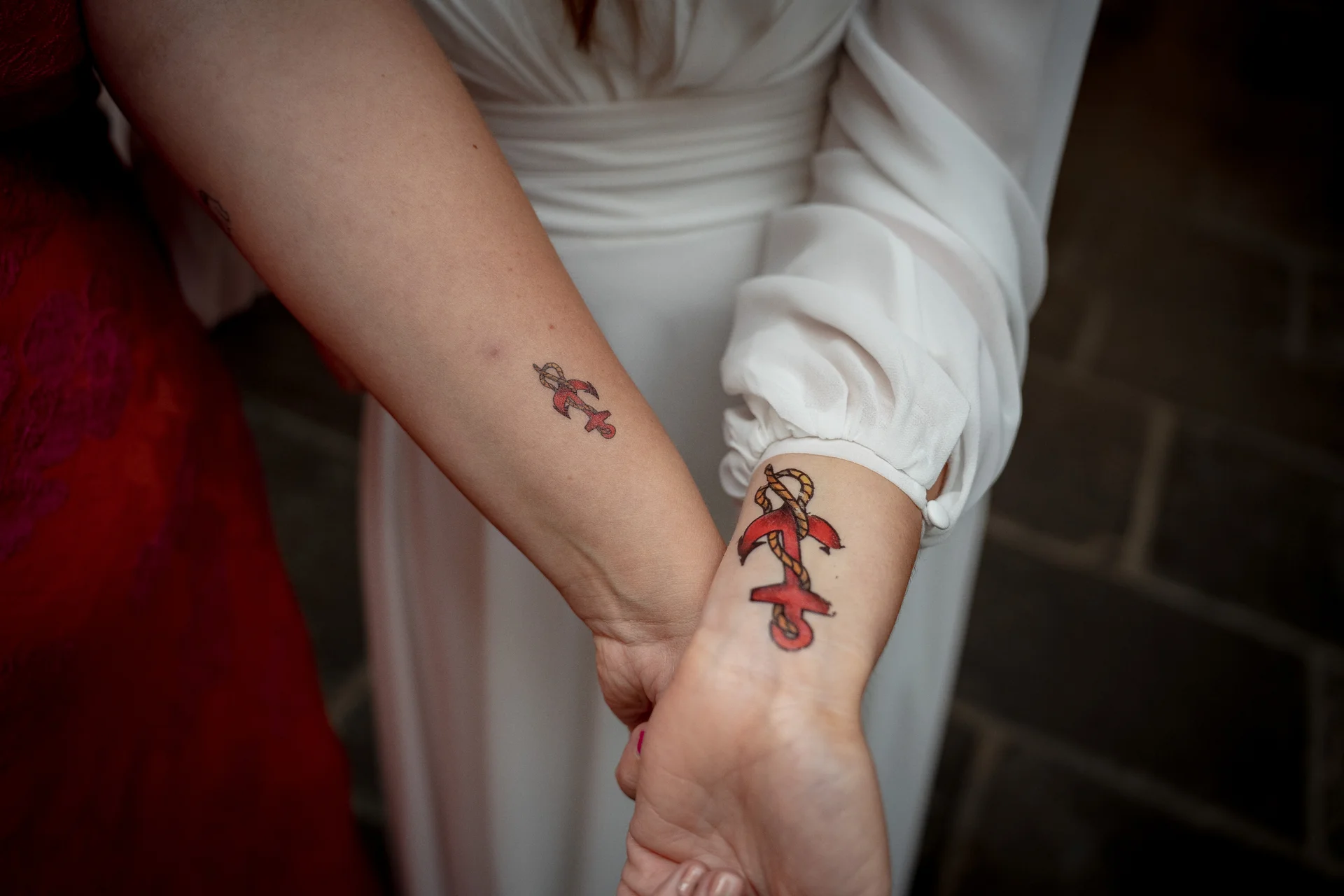 Two people display matching anchor tattoos on their inner wrists while holding hands, with one wearing a white long-sleeved dress and the other in a red dress.