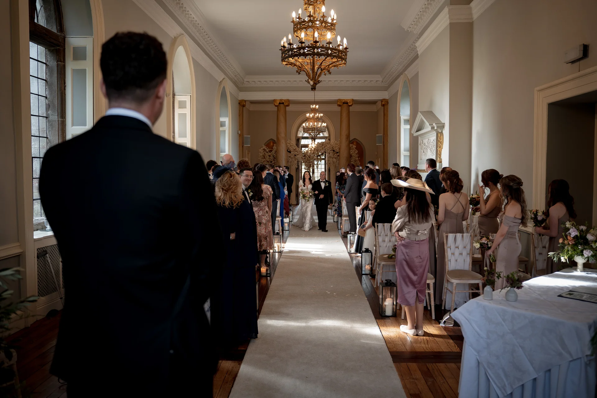 An over-the-shoulder shot captures the groom standing at the front of a grand, chandelier-lit hall while the bride walks down the aisle towards him.