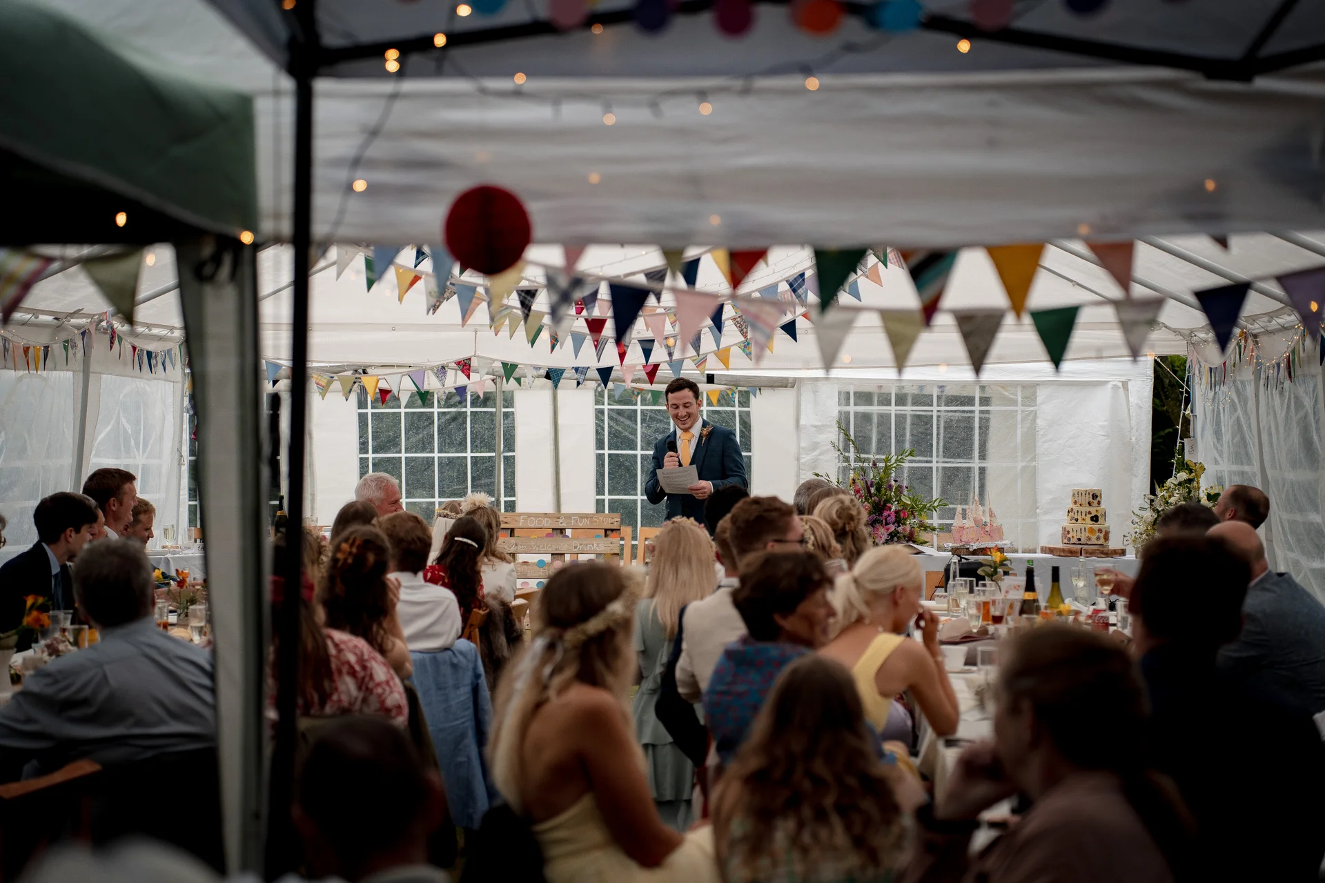 A groom delivers a speech inside a marquee decorated with colourful bunting and fairy lights while wedding guests listen attentively at tables.
