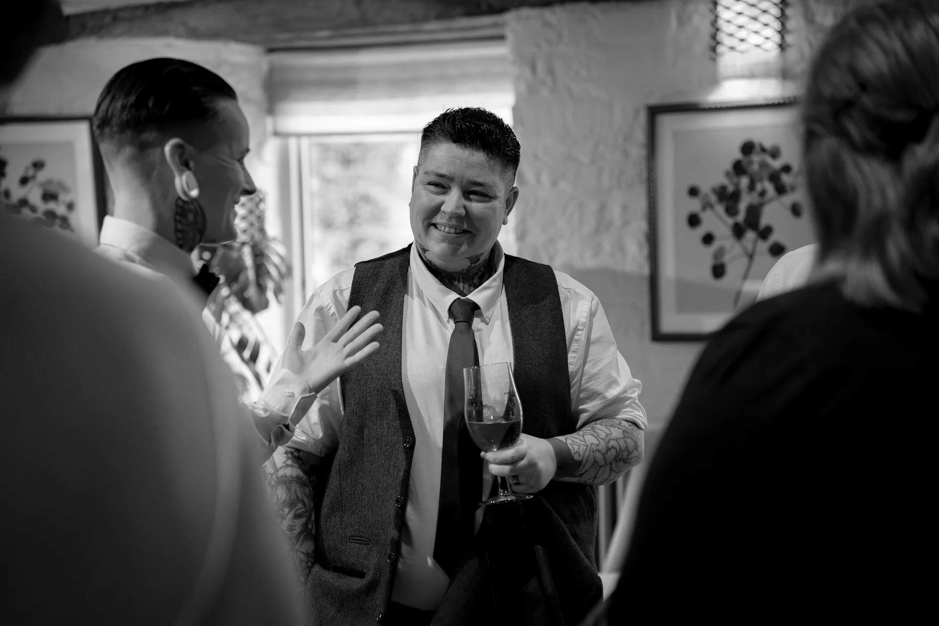 A smiling person in a waistcoat and tie holding a glass of wine talks and laughs with guests during a wedding reception.