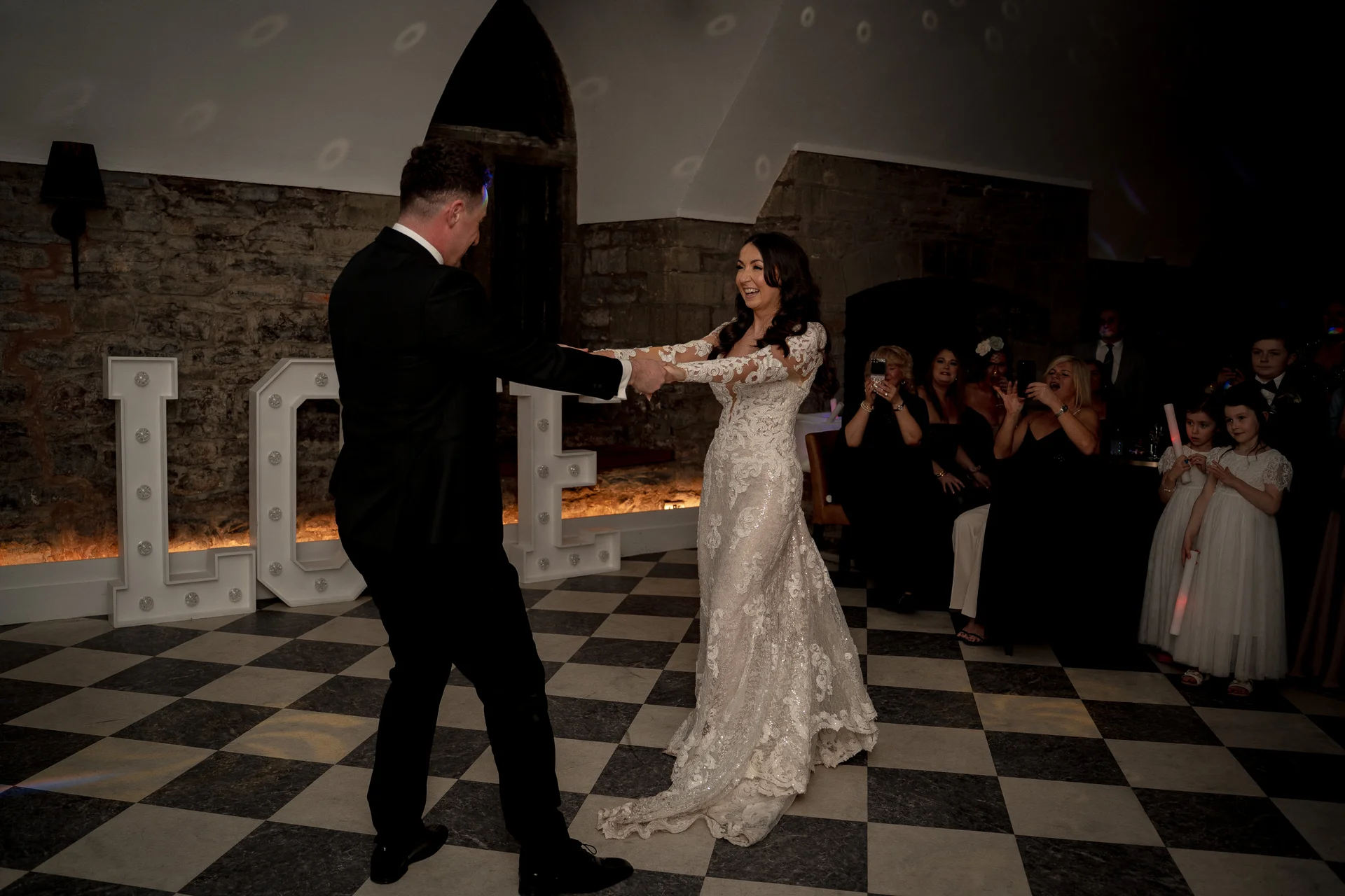 The newlywed couple share their first dance on a black and white checkered floor in a stone venue with illuminated letters in the background.