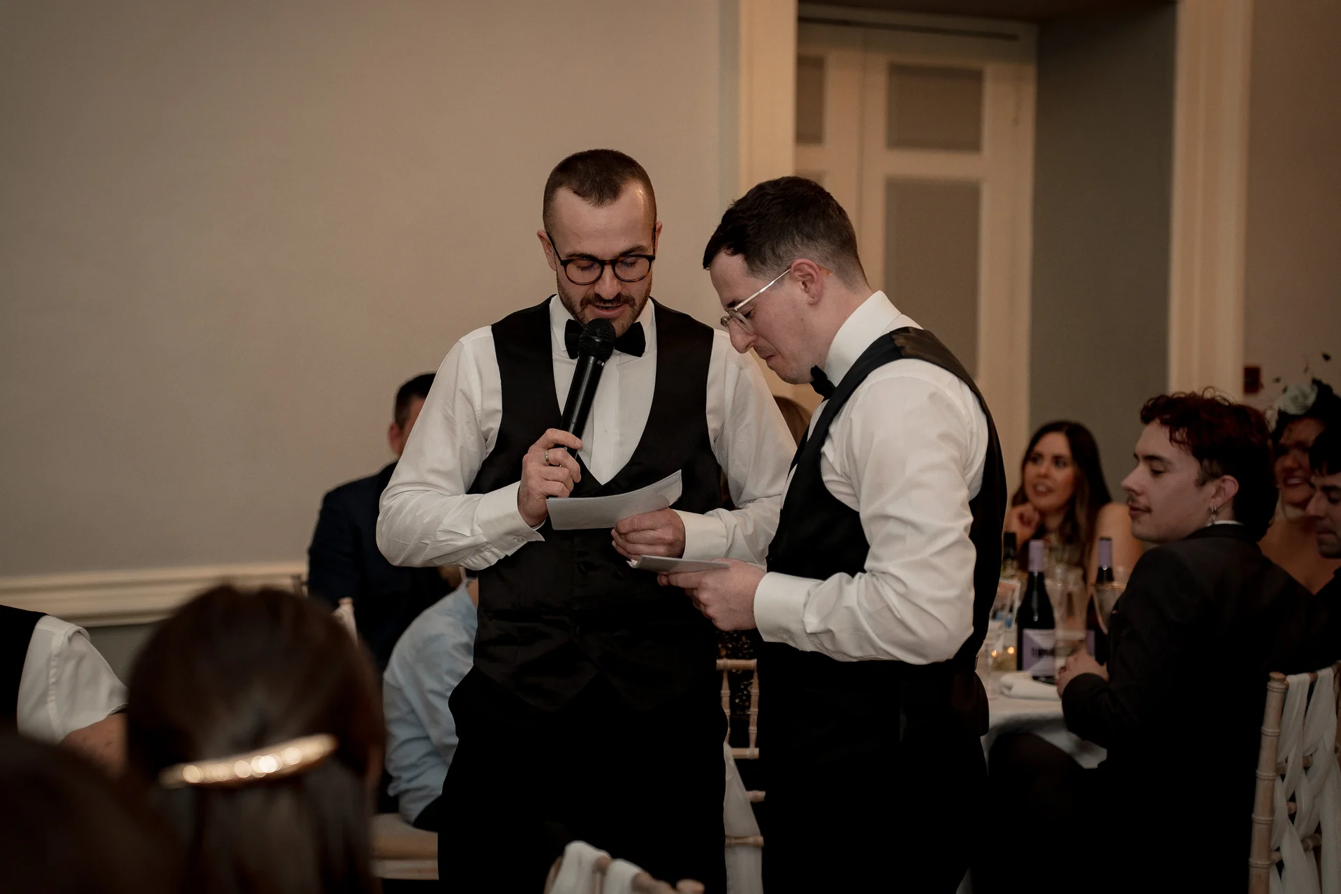Two men in waistcoats and bow ties deliver a speech at a wedding reception while guests watch from surrounding tables.