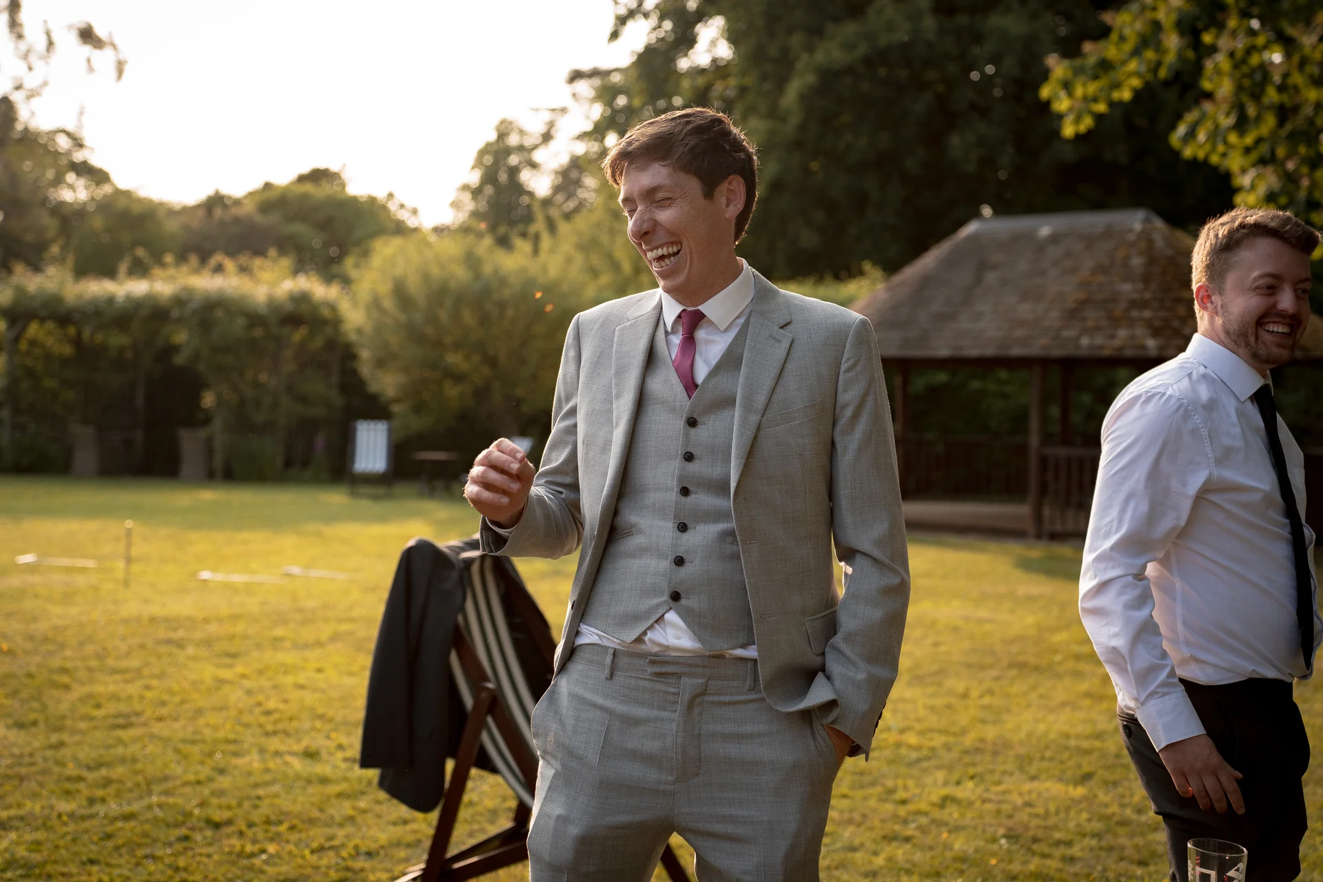A groom in a light grey three-piece suit and pink tie laughs heartily outdoors on a sunlit lawn during a summer wedding reception.