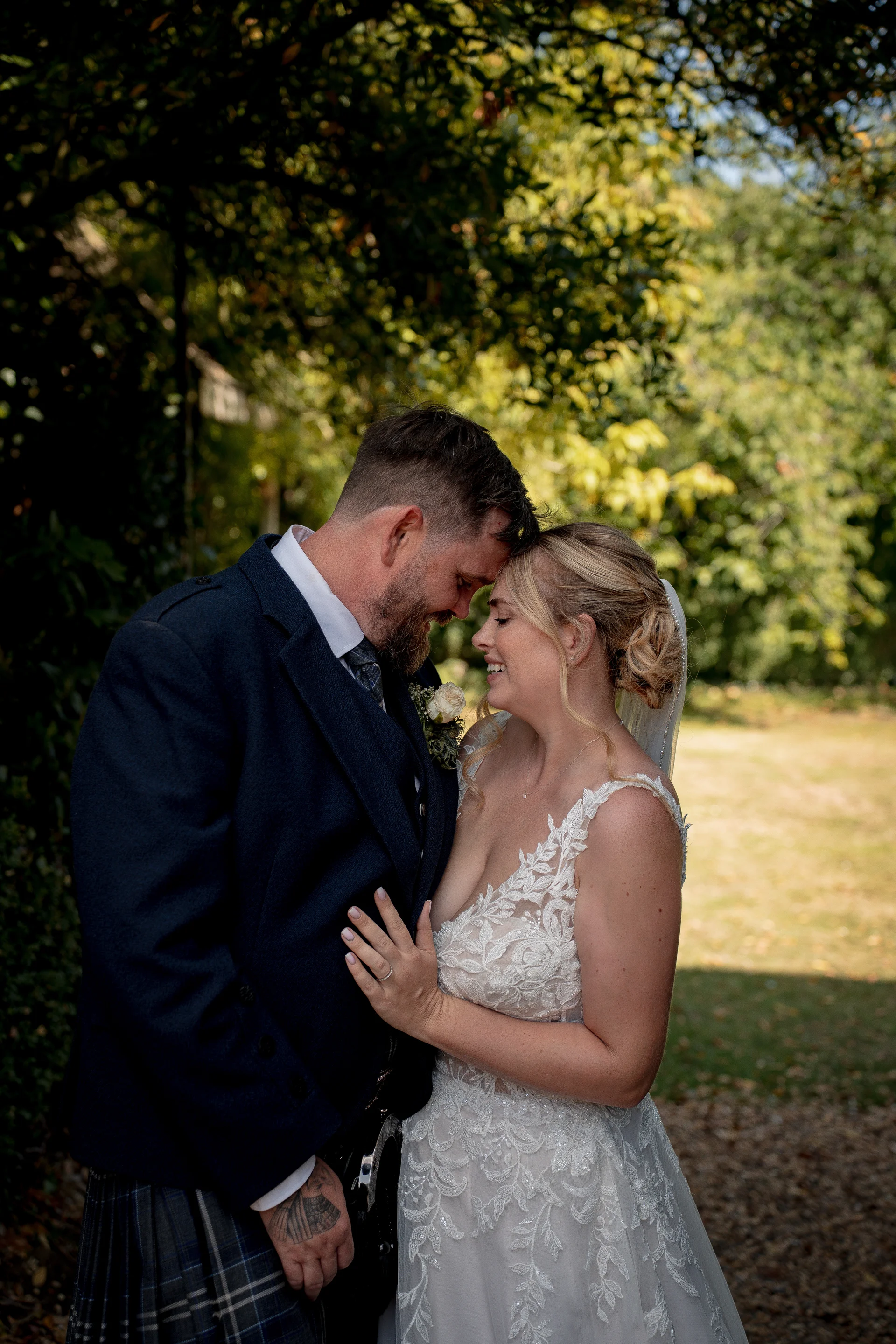A smiling bride and groom share a tender forehead touch outdoors amidst lush green foliage and dappled sunlight.