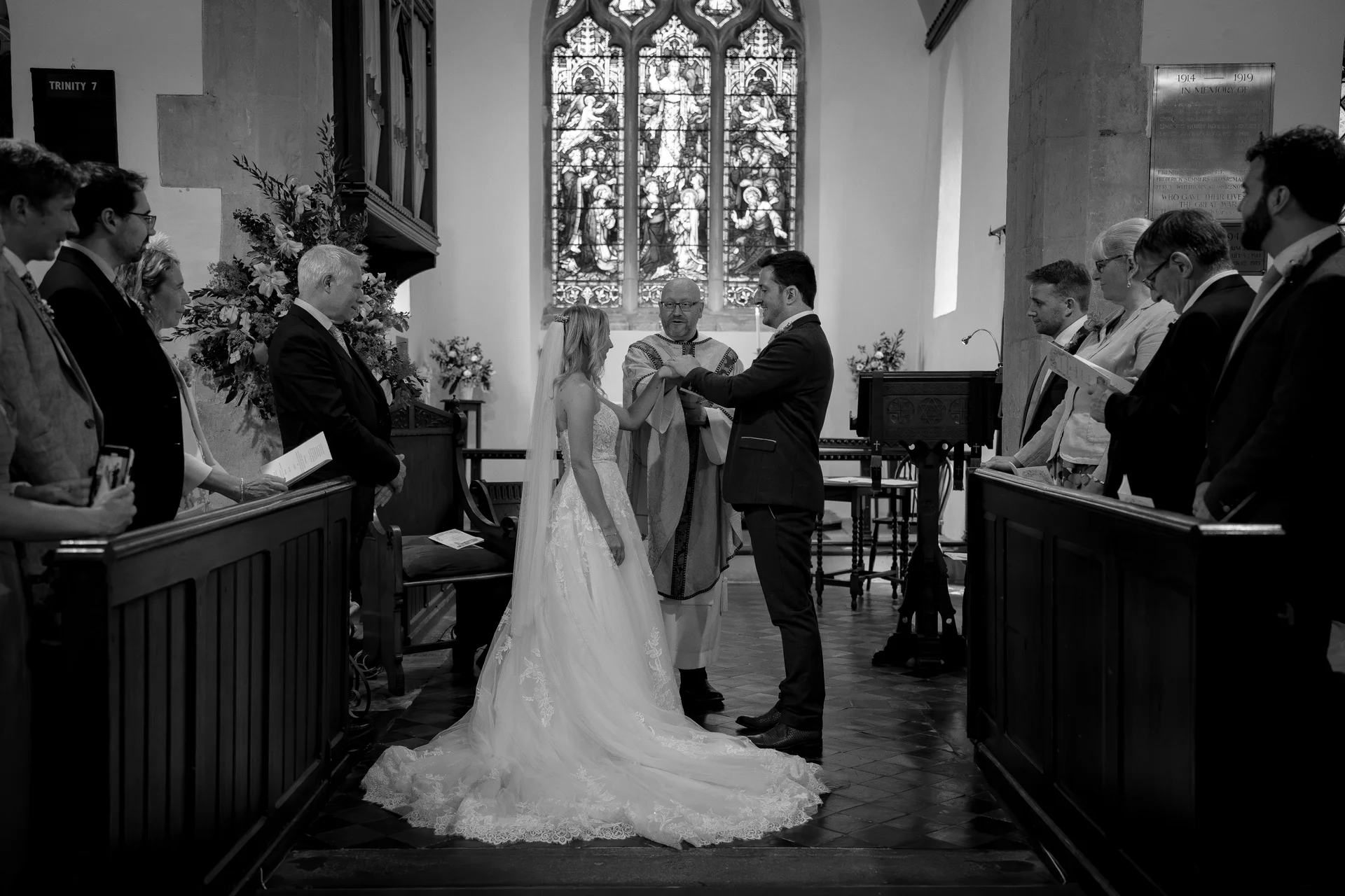 A black and white photograph captures the bride and groom exchanging rings at the altar of a traditional church with stained glass windows.