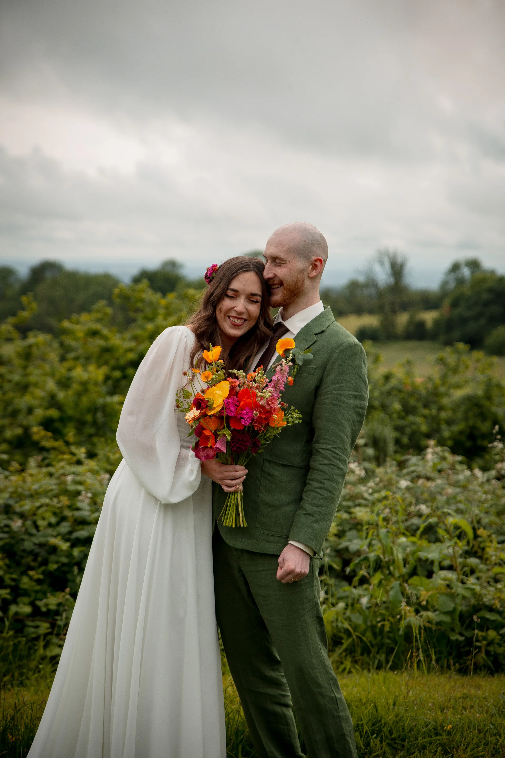A newlywed couple smiling affectionately outdoors against a moody overcast sky, holding a vibrant, colourful bouquet featuring orange and pink wildflowers.
